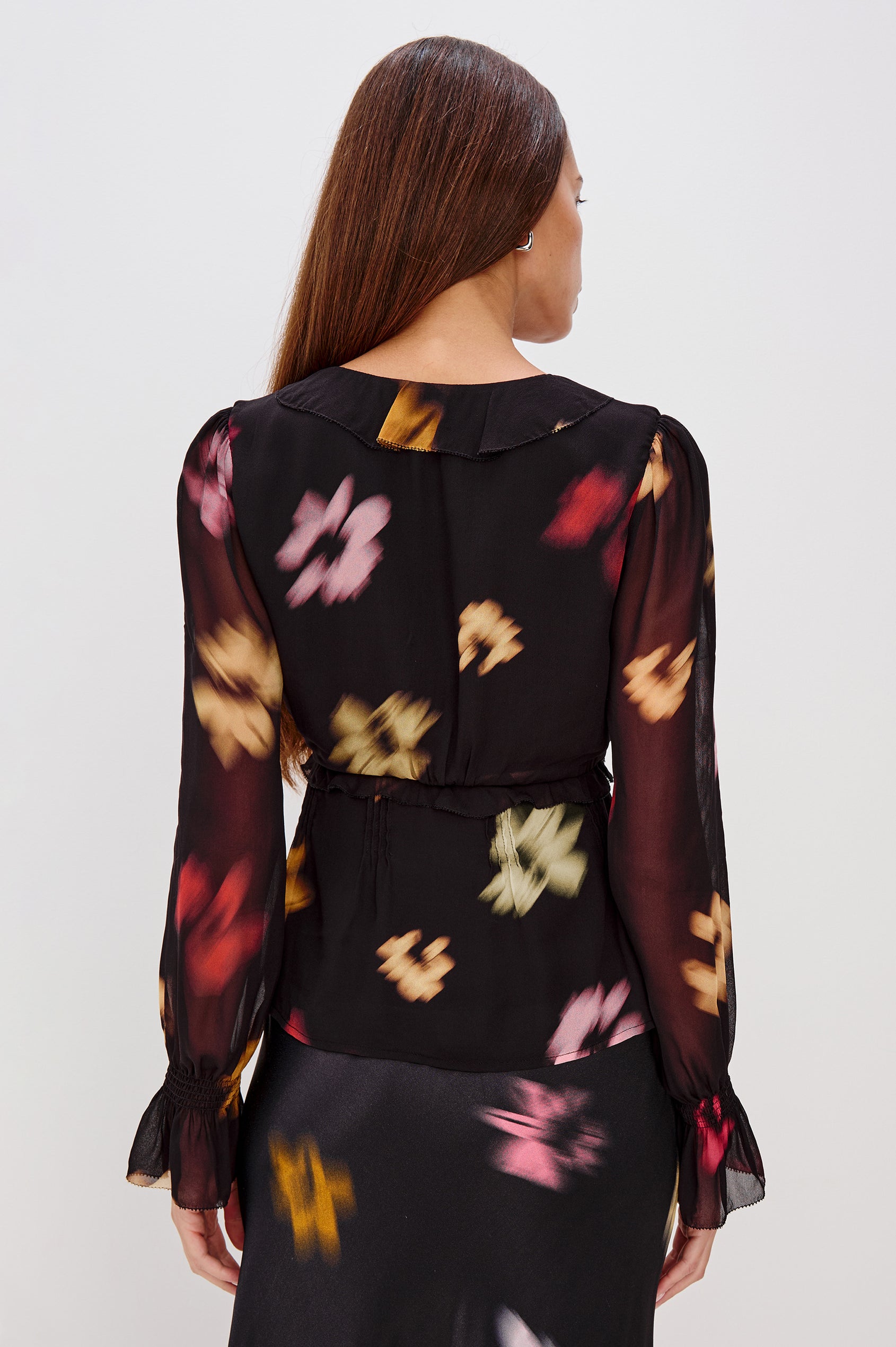 A lightweight chiffon blouse with a blurred floral print. The top features a deep V-neckline with cascading ruffles, button-front closures, and long sleeves finished with smocked bell cuffs. Back View Featured on Model.