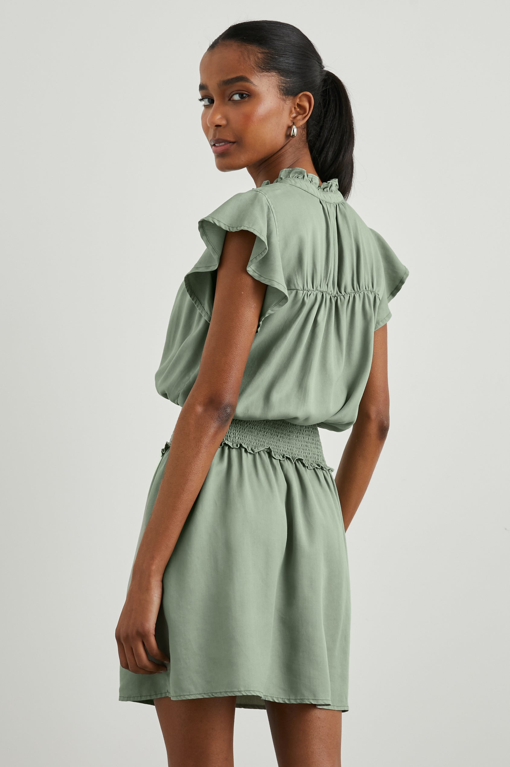 Amina Short Sleeve Mini Dress with Flutter Sleeves, a Ruffled Collar with Ruching, a Button Placket, and a Shirred Waist in Sage Denim Colorway - Back View Featured on Model 