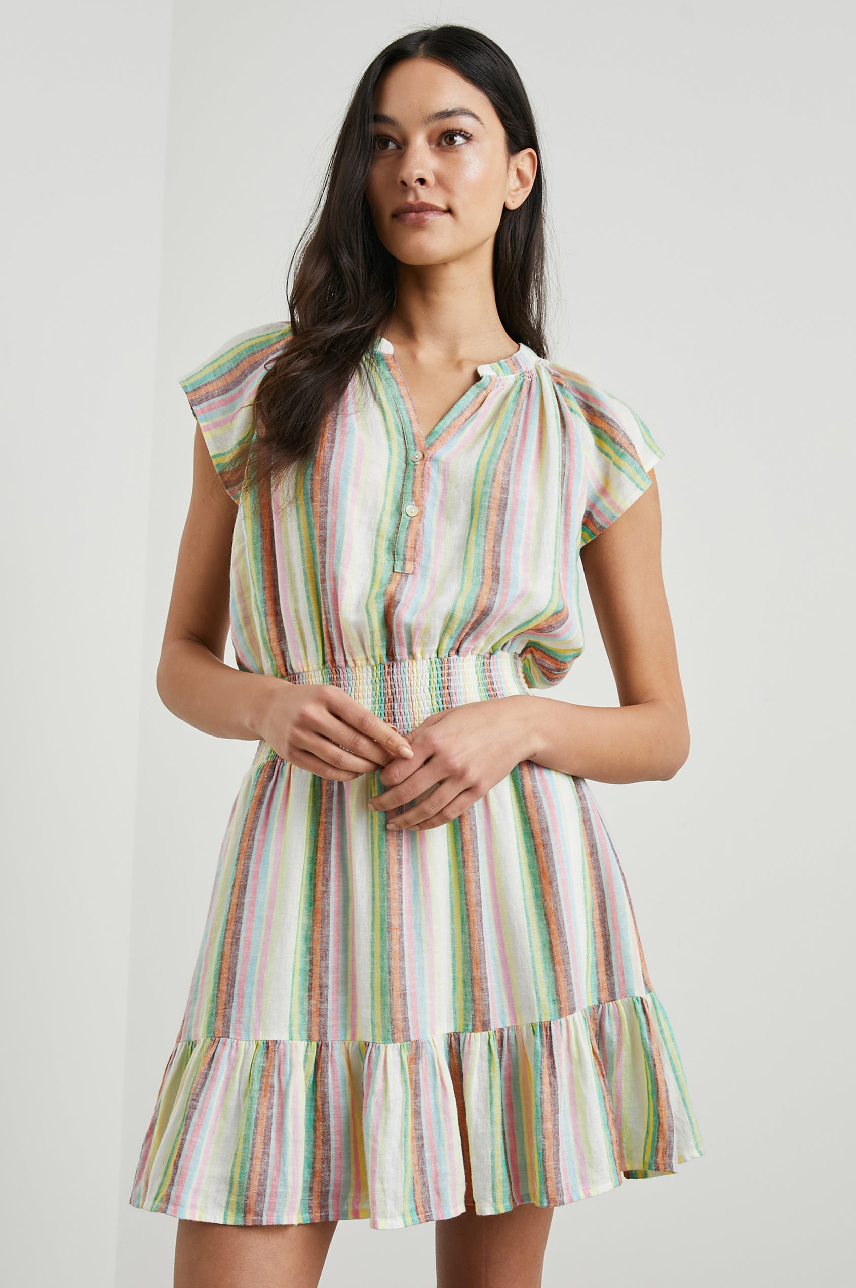  Augustine Short Sleeve Mini Dress with Flutter Cap Sleeves, a V Neck, a Smocked Waist, a Ruffled Hem, and a Placket with Buttons in Palais Stripe Colorway, Vertical Stripes in Green, Yellow, Orange, Brown, Pink, Blue and white - Front View Featured on Model 