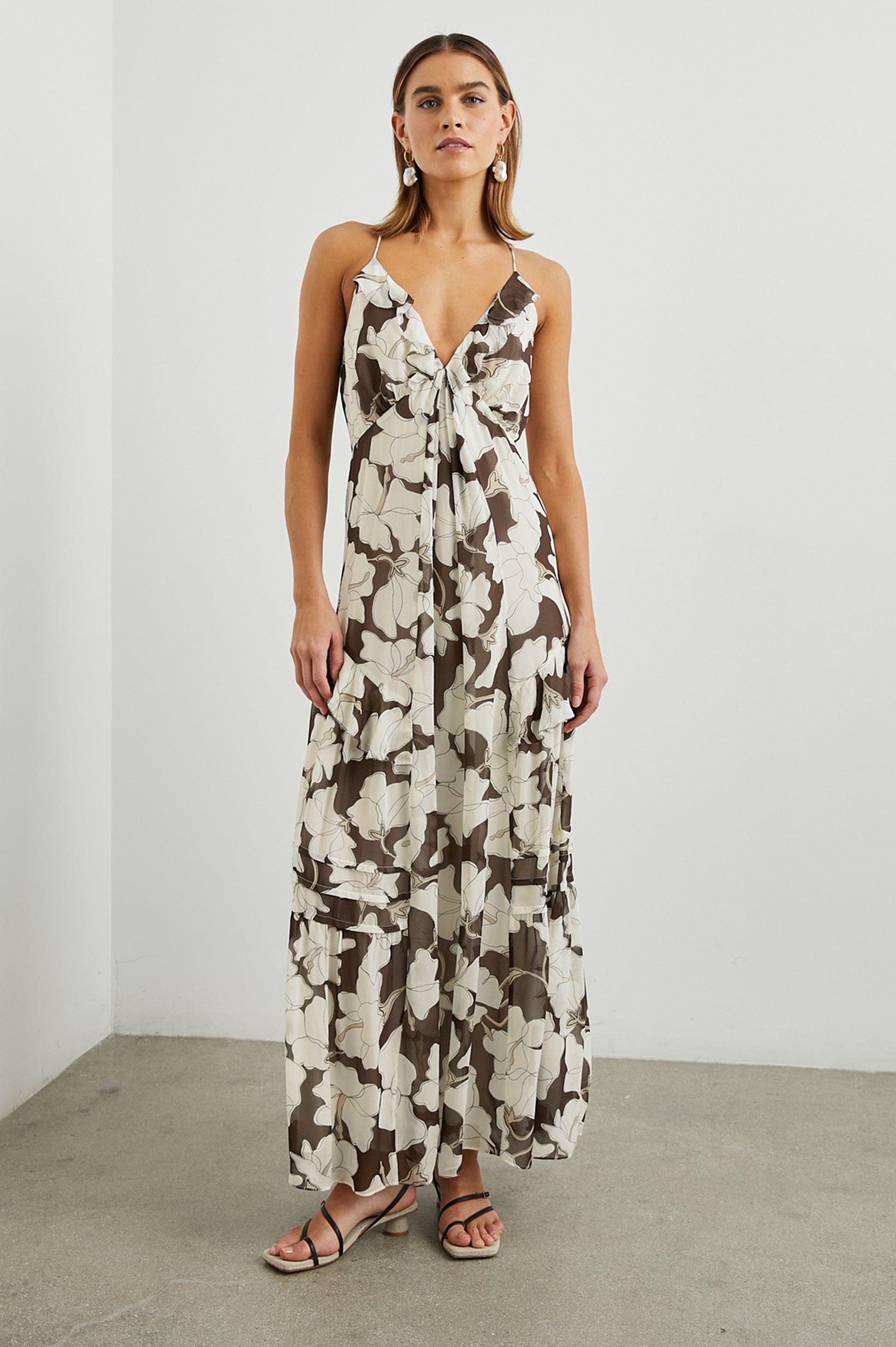Boa Sleeveless Maxi Dress with Thin Straps, a Deep V Neck, Empire Waist, Frills along the Neckline and Skirt and a Back Neck Tie in Mocha Floral Colorway, with large Cream Flowers on Mocha - Front Full Body View Featured on Model