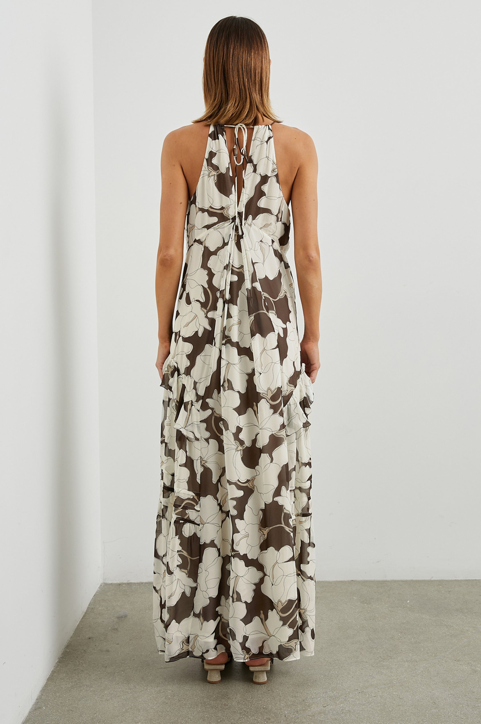 Boa Sleeveless Maxi Dress with Thin Straps, a Deep V Neck, Empire Waist, Frills along the Neckline and Skirt and a Back Neck Tie in Mocha Floral Colorway, with large Cream Flowers on Mocha - Back Full Body View Featured on Model
