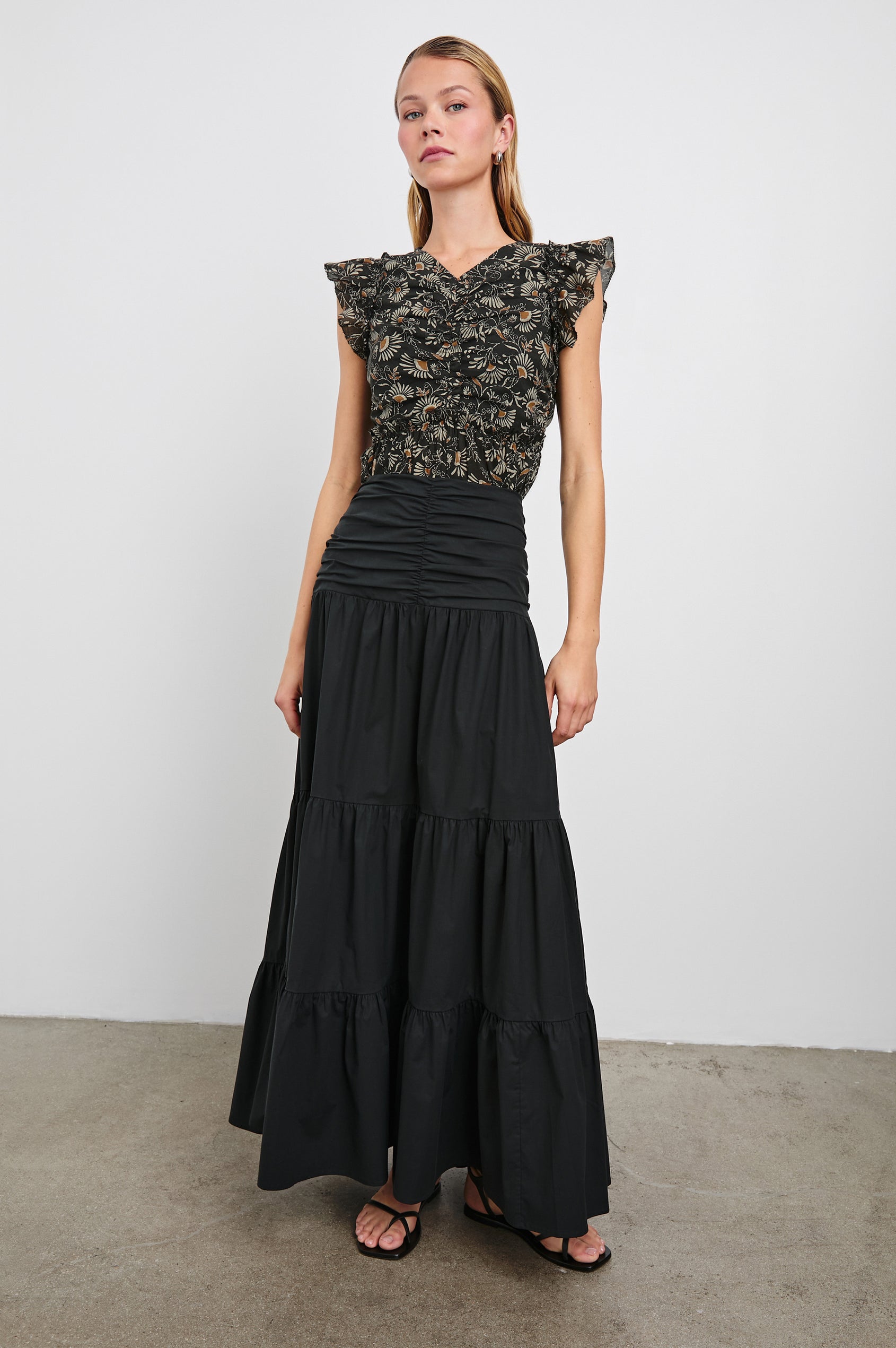 Cali Top with Flutter Sleeves, a V Neckline, Horizontal pleats along the bodice and vertical pleats at the hem in Cassia Colorway, with white and orange flowers on black - Front Full Body View Featured on Model