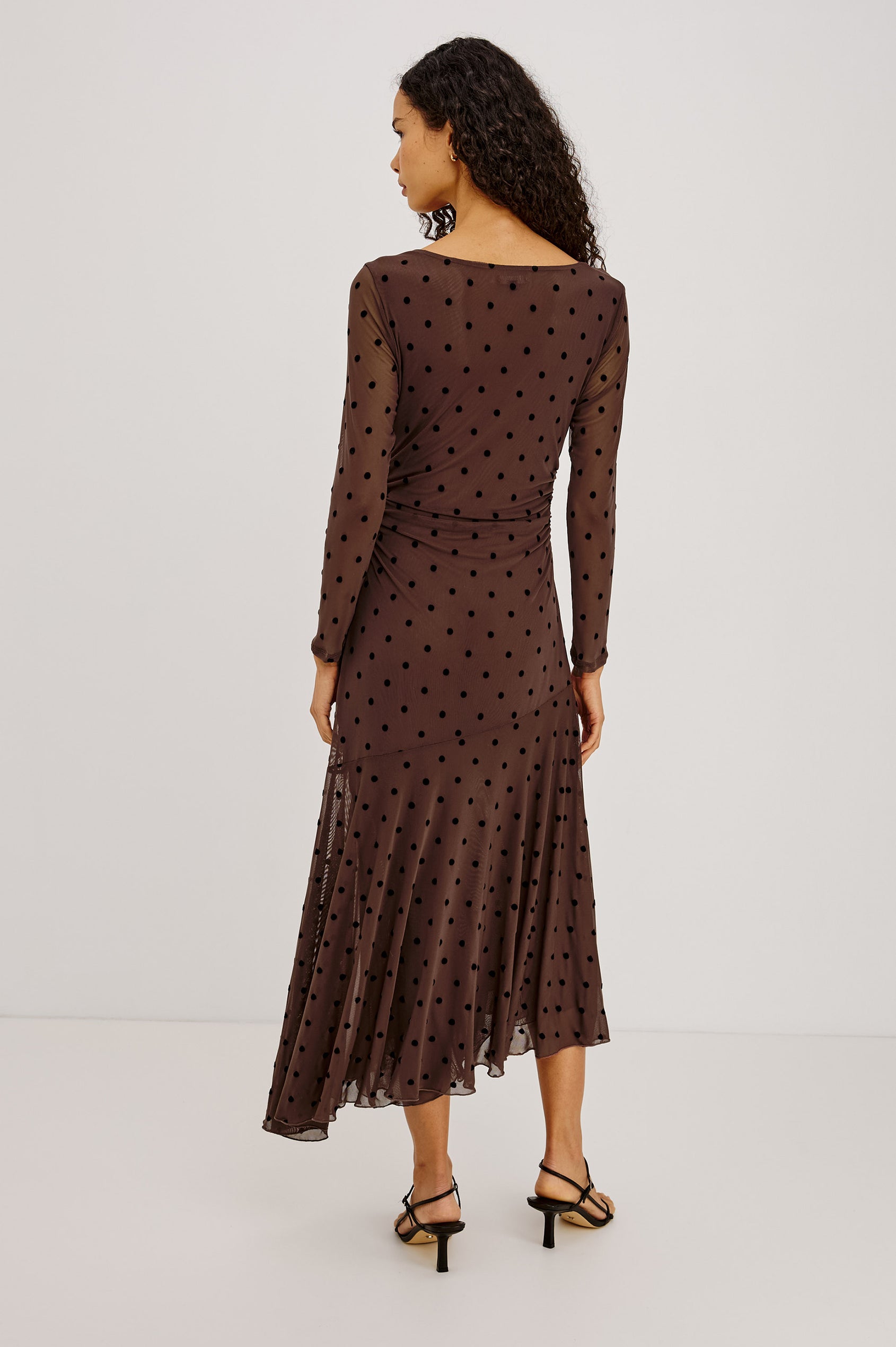 Cambria Midi Dress in Semi-Sheer Stretch Mesh With Velvet Polka Dots, Self-Lined Body, Asymmetric Cowl Neckline, Ruched Side Waist, and Softly Flared Skirt in Chocolate Polka Dot Colorway - Full Body Back View Featured on Model
