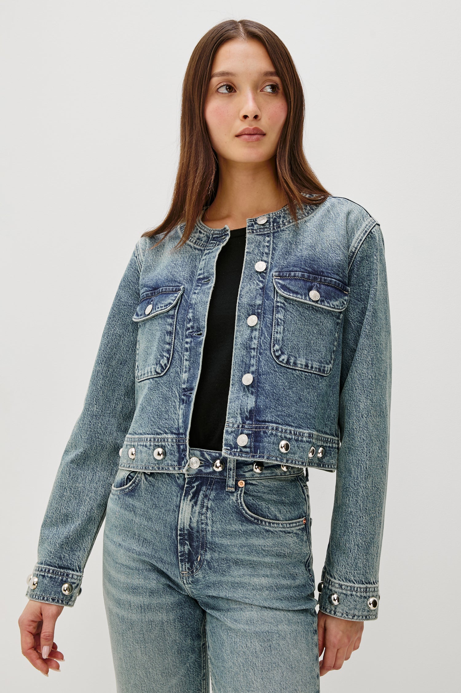 The Capistrano is a cropped denim jacket with a scoop neck, it has a button front, two chest pockets and silver studs on the sleeve cuffs and hem in Desert Sky colorway. Front view featured on model.