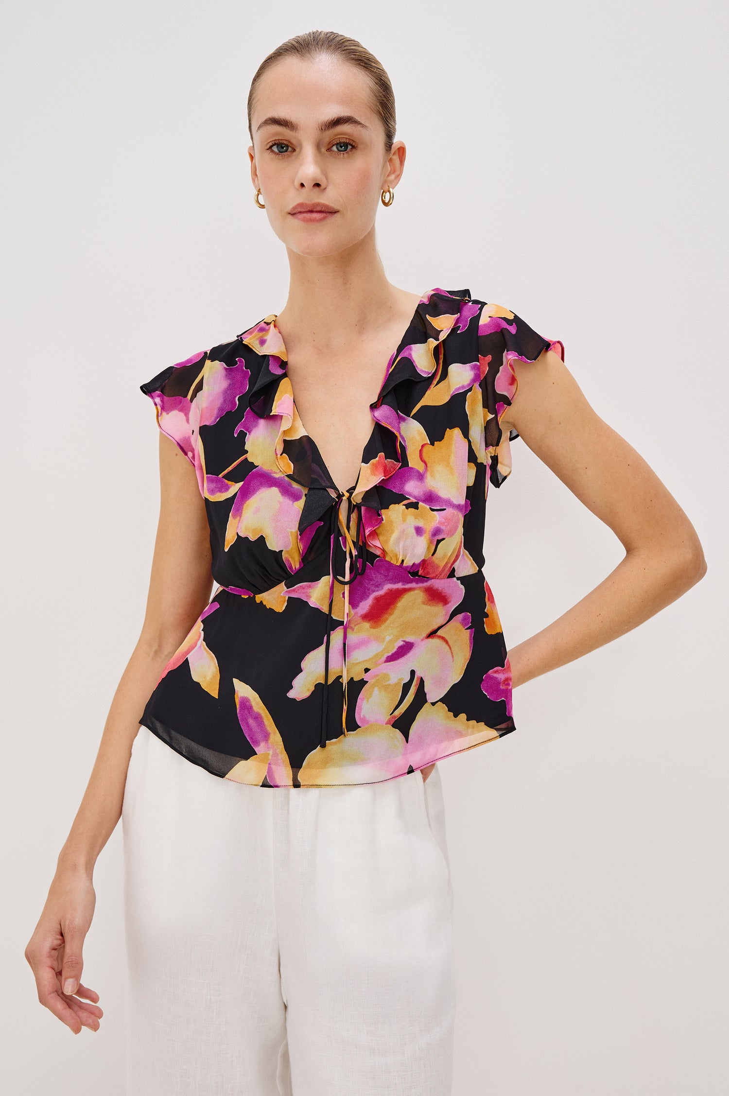 Carmine blouse, a chiffon top with ruffled cap sleeves, ruffles at the v-neckline, a front tie, and empire waist silhouette in Dusk Orchid Colorway. Front View Shown on Model.