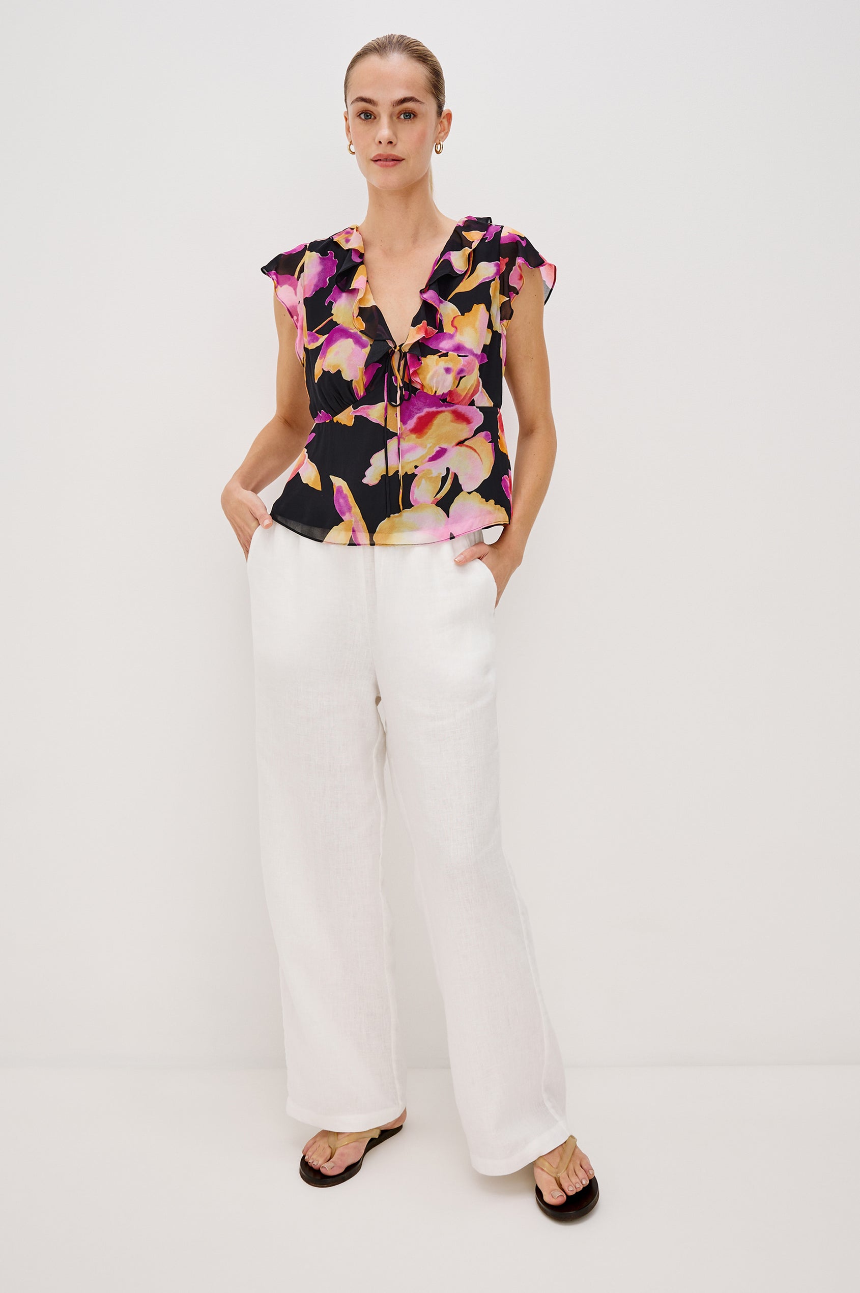 Carmine blouse, a chiffon top with ruffled cap sleeves, ruffles at the v-neckline, a front tie, and empire waist silhouette in Dusk Orchid Colorway. Full Body Front View Shown on Model.