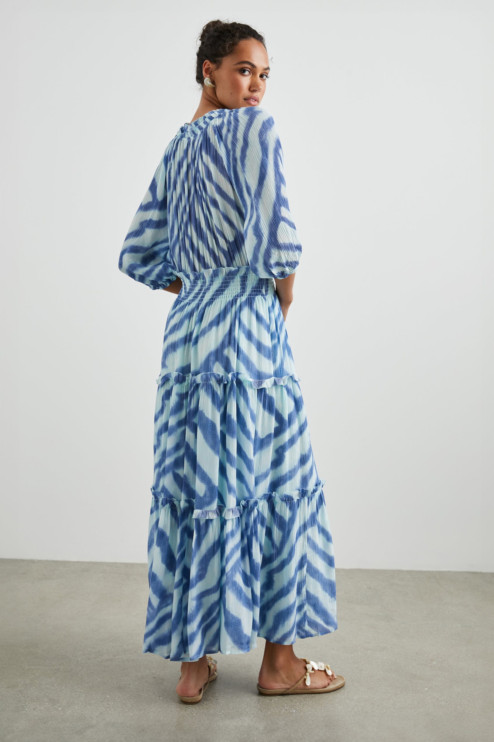 Caterine Maxi Dress with Mid-length Sleeves, a V neck, a Shirred Collar with a tie, a Smocked Waist, and a Tiered Skirt in Blue Watercolor Stripes Colorway, a pattern with blue on light blue - Back Full Body View Featured on Model