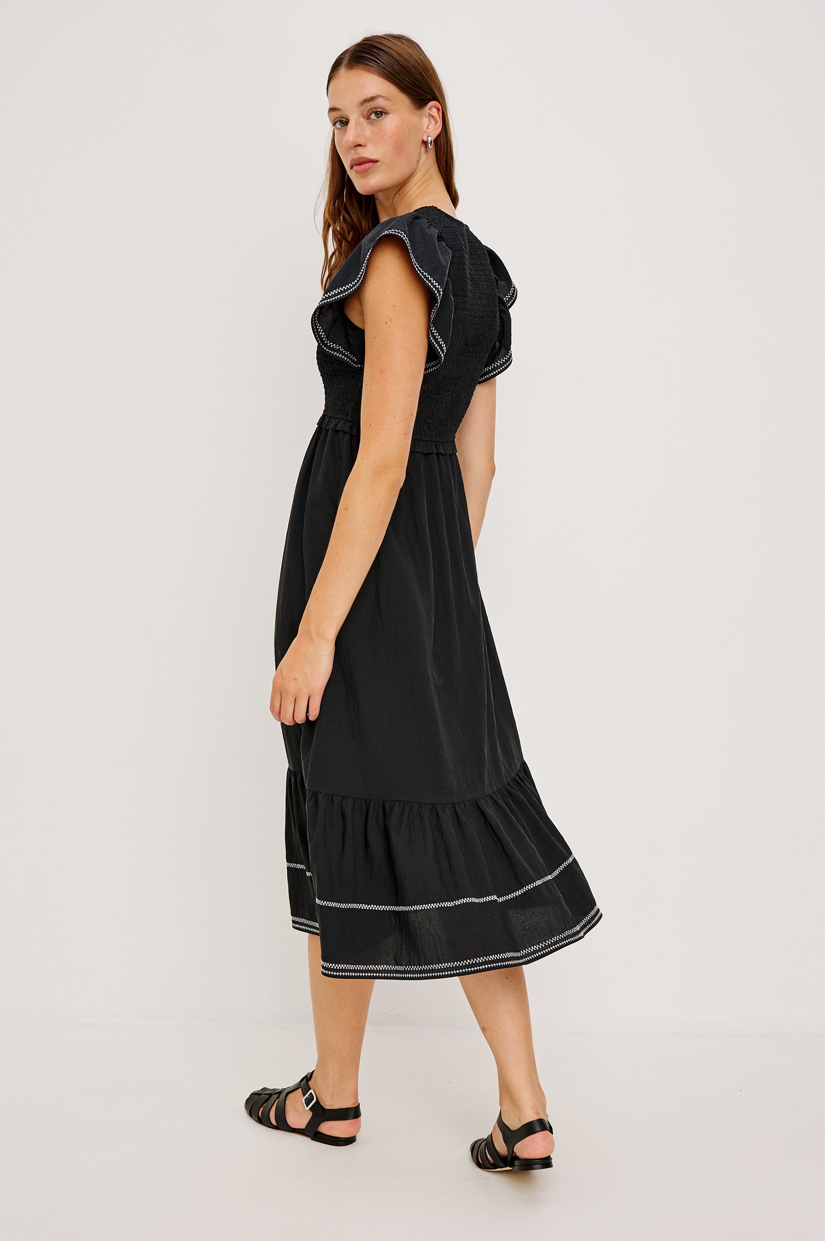 Clementine Midi Dress with a Deep V Neckline, Flutter Sleeves, Smocked Top, Two Side Pockets and contrast white stitching on the Sleeves and Hem in Black Colorway- Back Full Body View Featured on Model.