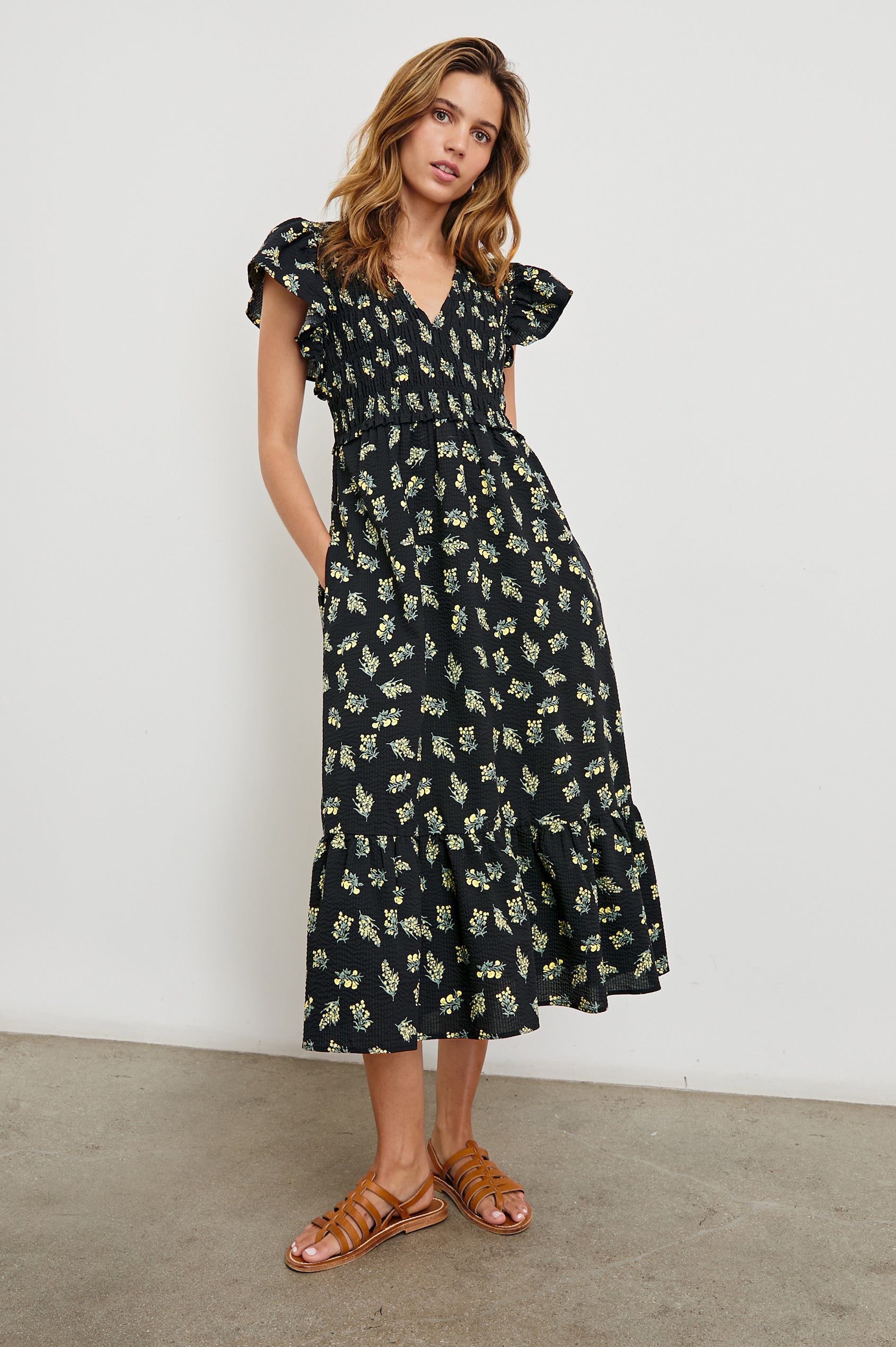 Clementine Midi Dress with a Deep V Neckline, Flutter Sleeves, Smocked Top and Two Side Pockets in Dark Amaryllis Colorway, Yellow and Green flowers on Black - Front Full Body View Featured on Model