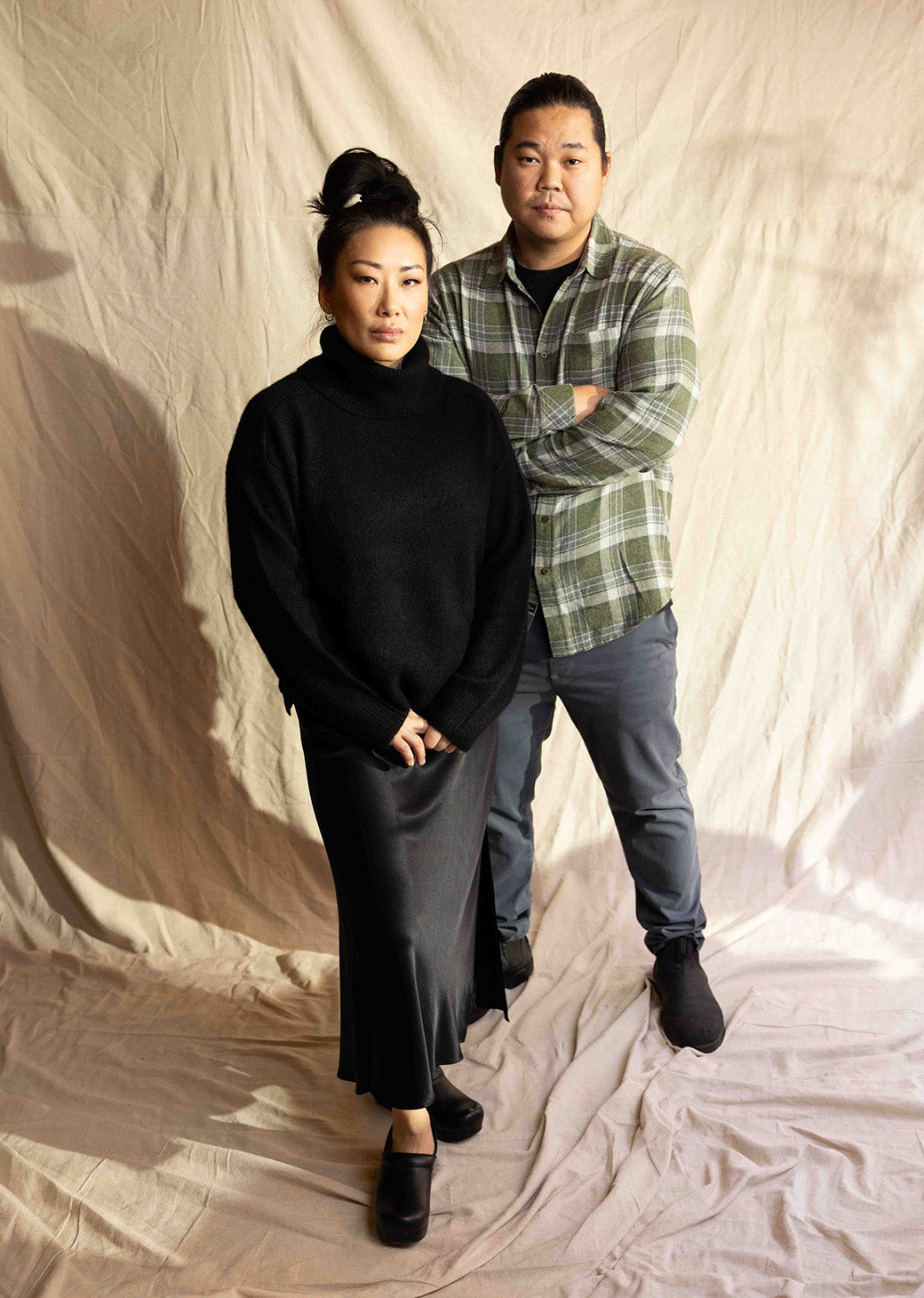 MALE AND FEMALE. FEMALE IS WEARING ESTELLE SWEATER (LONG SLEEVE TURTLENECK CASHMERE SWEATER) IN BLACK AND ANYA SKIRT (MID LENGTH SATIN SKIRT) IN BLACK. MALE IS WEARING LENNOX SHIRT (LONG SLEEVE BUTTON UP) IN GREEN PLAID.