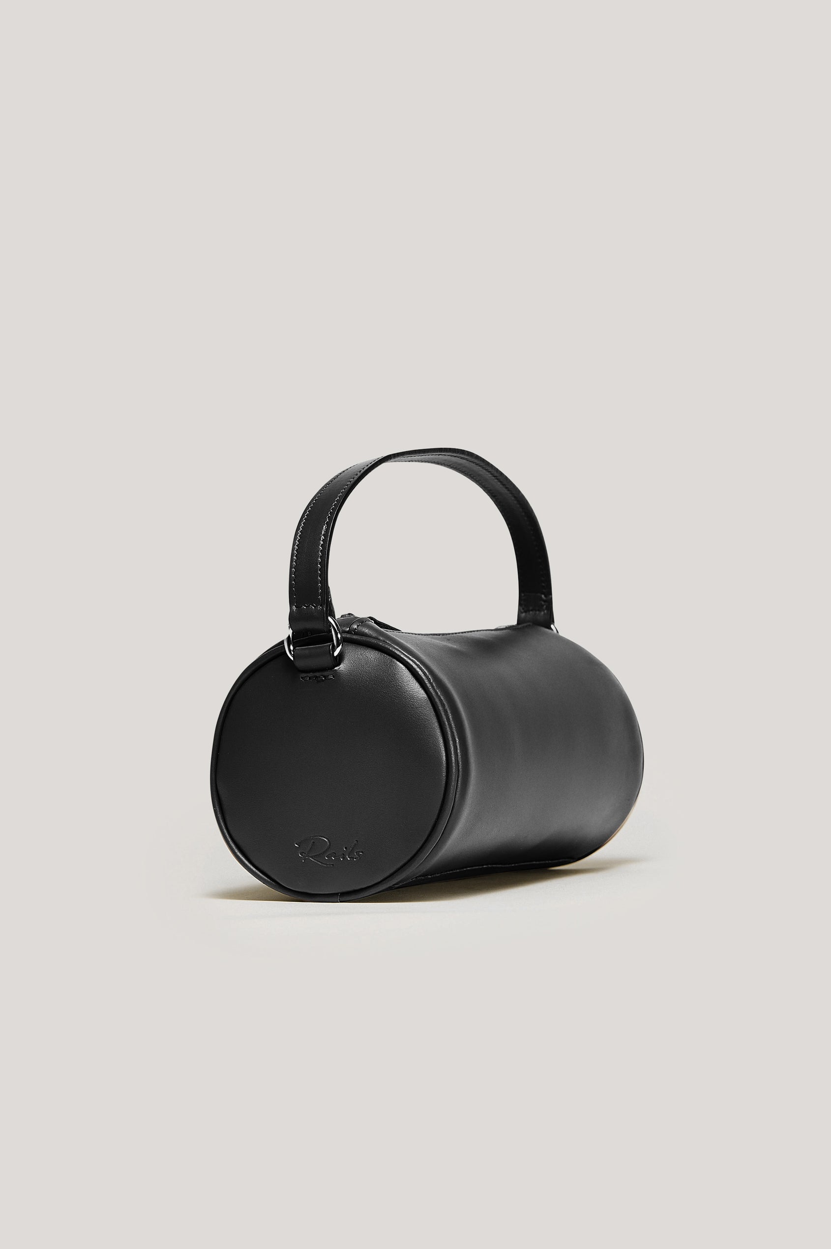 The Como is a mini barrel bag, made from smooth leather with a fixed top strap, a removable and adjustable buckled crossbody strap, a top zip, a side-embossed logo, and a branded interior label, in Black Leather colorway. Side angled view featured.
