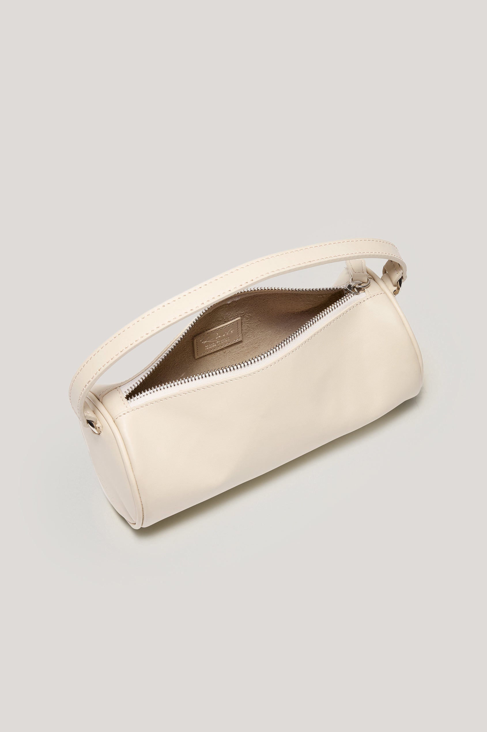 The Como is a mini barrel bag, made from smooth leather with a fixed top strap, a removable and adjustable buckled crossbody strap, a top zip, a side-embossed logo, and a branded interior label, in Bone Leather colorway. Top view of interior featured.