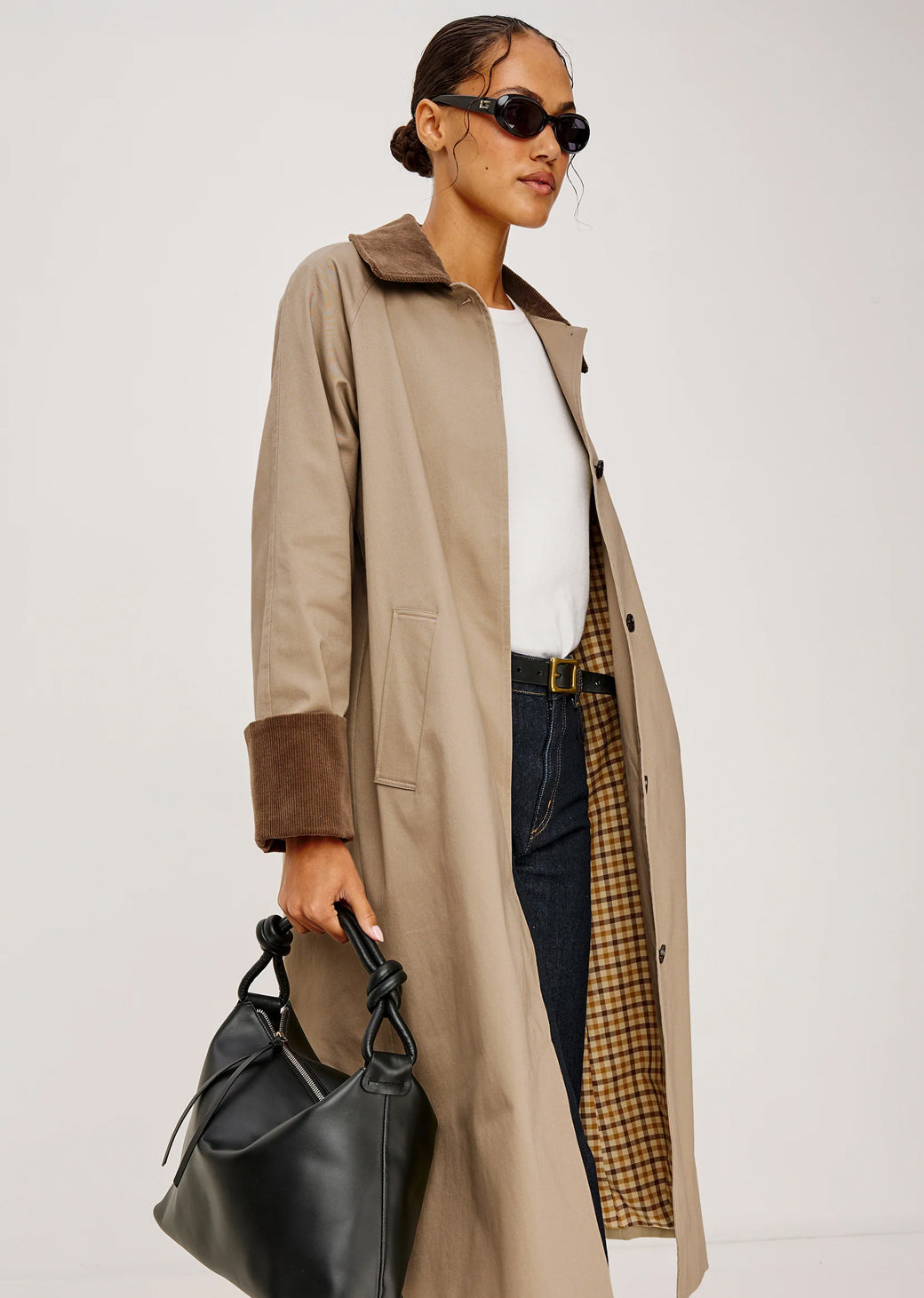 Woman wearing a beige trench coat holding a black handbag on a white background.