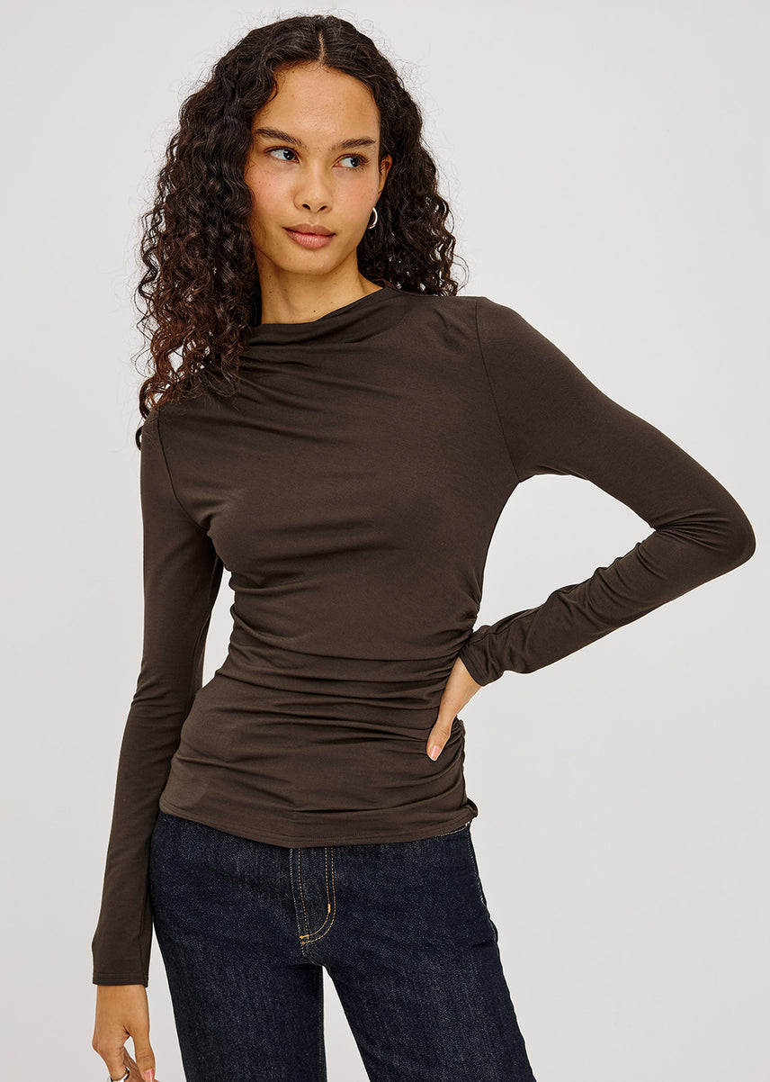 A woman modeling a dark brown long-sleeve ruched top with a draped neckline, paired with dark blue jeans, against a light gray studio background.