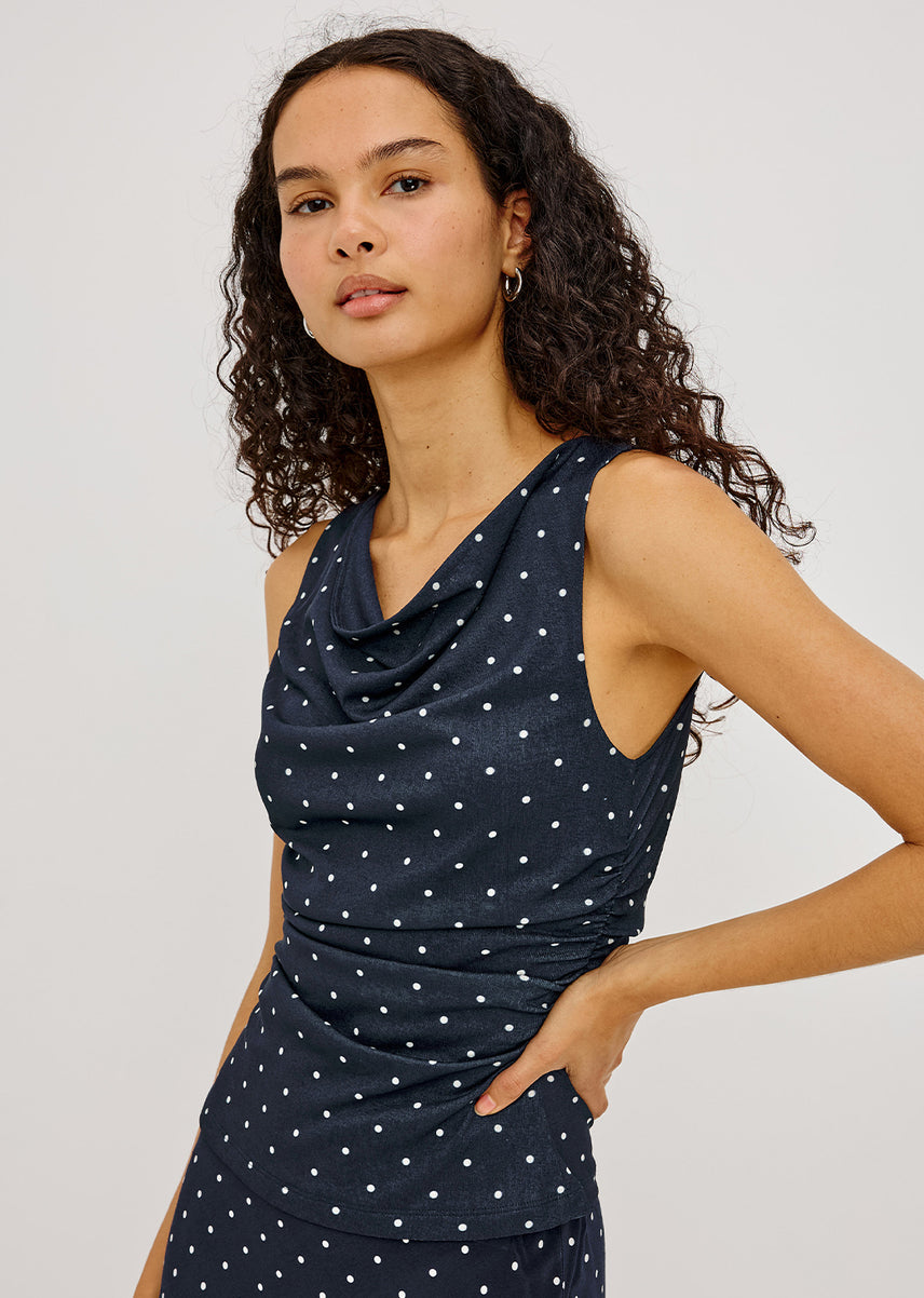 A woman modeling a sleeveless navy blue dress with a white polka dot pattern, featuring a draped cowl neckline and ruched detailing at the waist, set against a plain light gray background.
