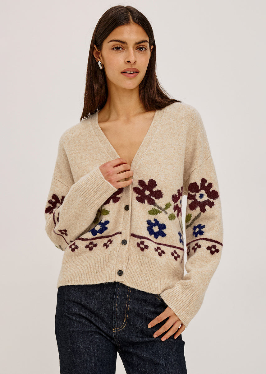 A woman modeling a cream-colored knit cardigan with colorful floral patterns in purple, blue, and green, paired with dark blue jeans against a plain light gray background.
