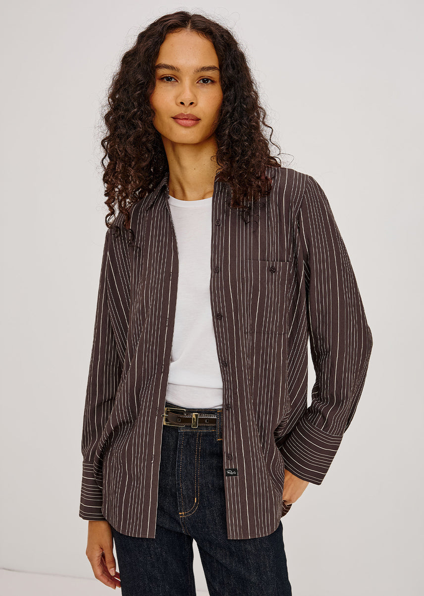 A woman with long curly dark hair models a brown vertically striped button-up shirt jacket over a white t-shirt and dark blue jeans, photographed against a light gray studio background.