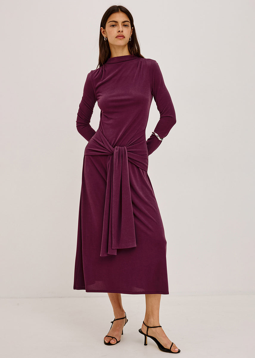 A woman modeling a long-sleeve midi dress in deep plum purple. The dress features a mock neck, gathered fabric at the waist with a matching fabric tie, and is styled with black strappy high-heeled sandals against a plain light gray studio background.