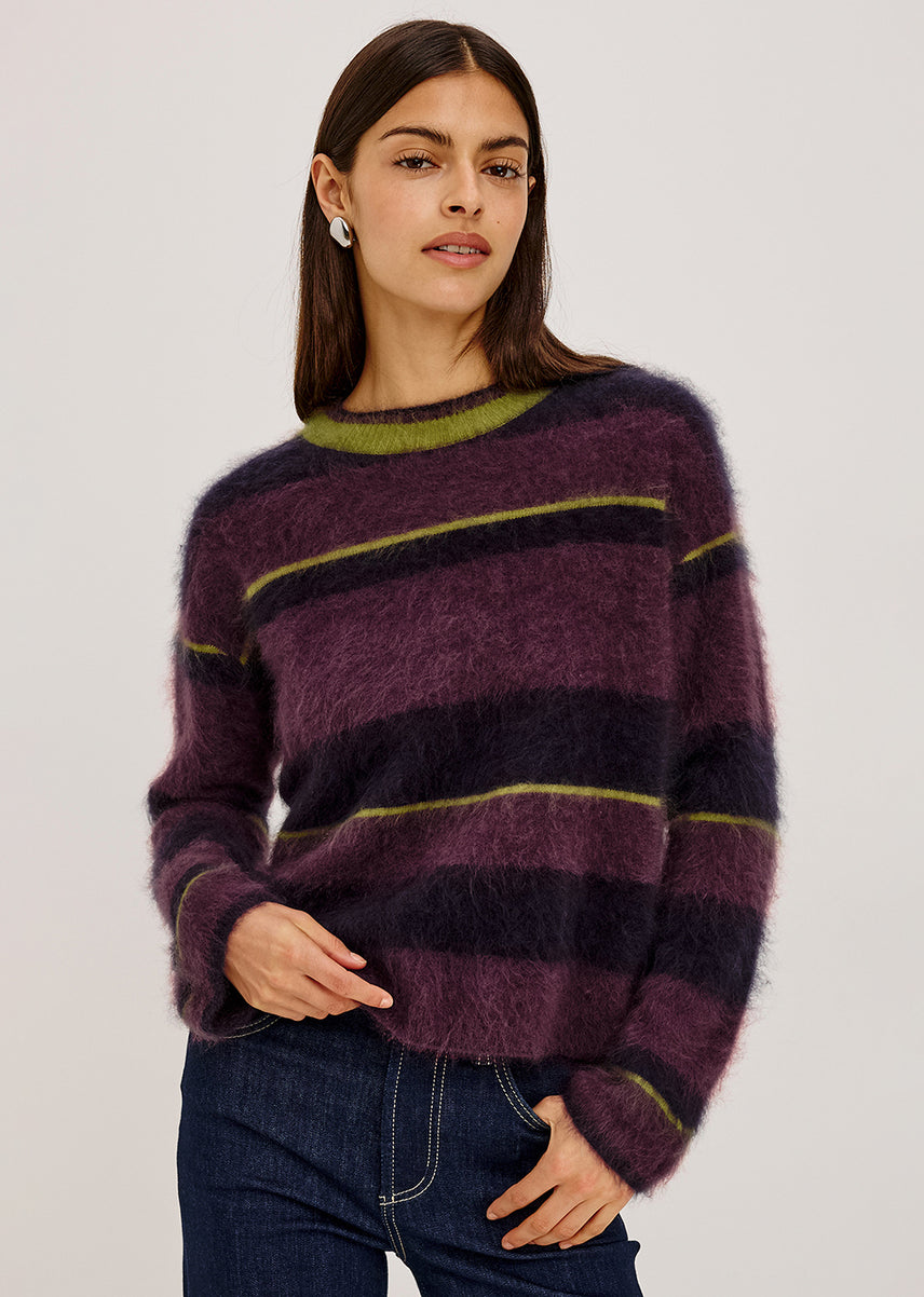 A woman modeling a fuzzy, long-sleeve striped sweater in shades of purple, dark navy, and olive green, paired with dark blue jeans against a plain light gray background.