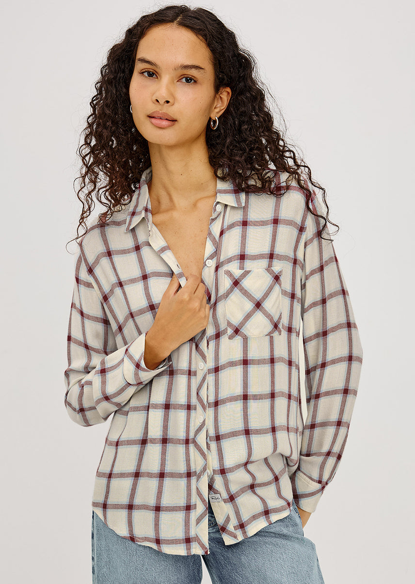 A woman with curly dark hair wearing a long-sleeve plaid button-up shirt in cream, white, and burgundy, paired with blue jeans, against a light gray studio background.