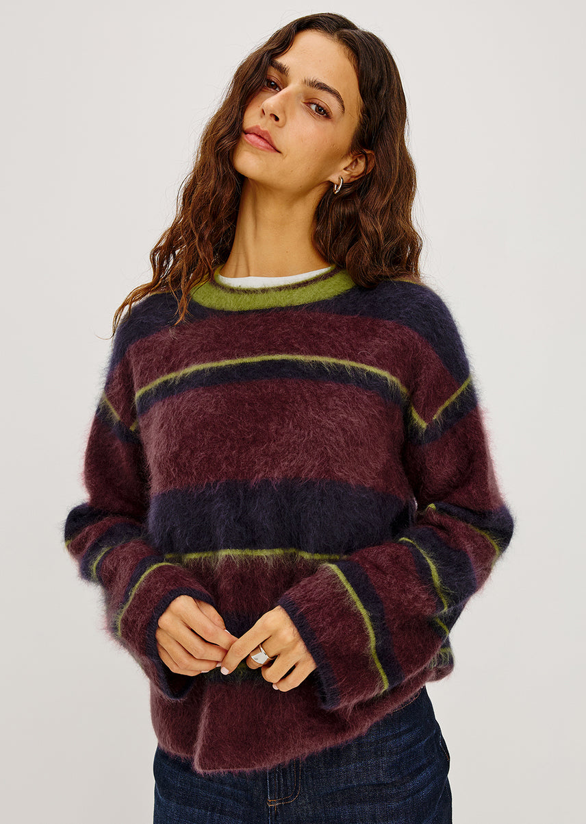 A woman modeling a fuzzy, long-sleeve striped sweater in shades of purple, dark navy, and olive green, paired with dark blue jeans against a plain light gray background.