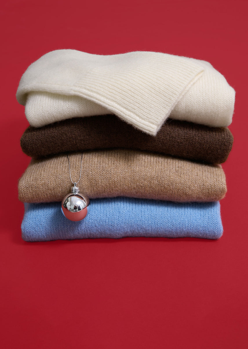 Stack of four folded knit sweaters in cream, dark brown, camel, and light blue, with a silver ornament hanging from a thin chain against a red background.
