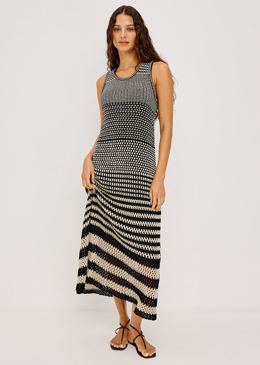 A woman modeling a sleeveless knit maxi dress with a black and beige horizontal striped pattern, photographed against a plain light gray studio background.