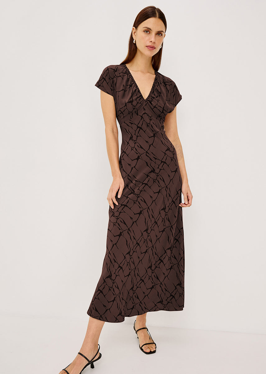 A woman modeling a dark brown midi dress with a black abstract pattern, short flutter sleeves, and a V-neckline, set against a plain light gray studio background.