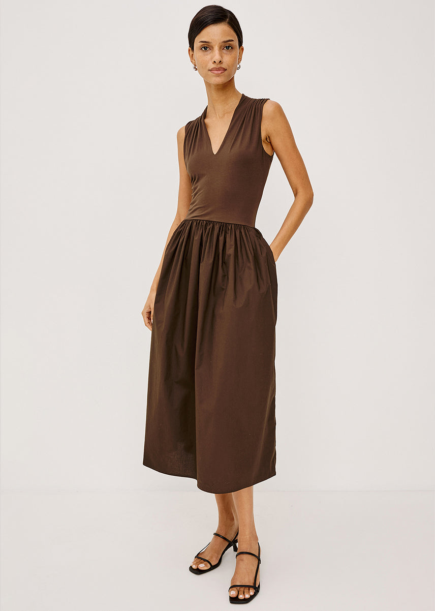 A woman modeling a sleeveless brown midi dress with a V-neckline and gathered waist, paired with black strappy sandals, against a plain light gray studio background.