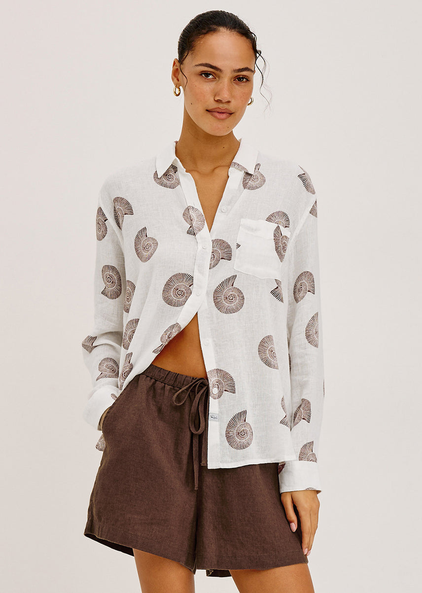 Woman wearing a white blouse with a pattern and brown shorts on a plain background