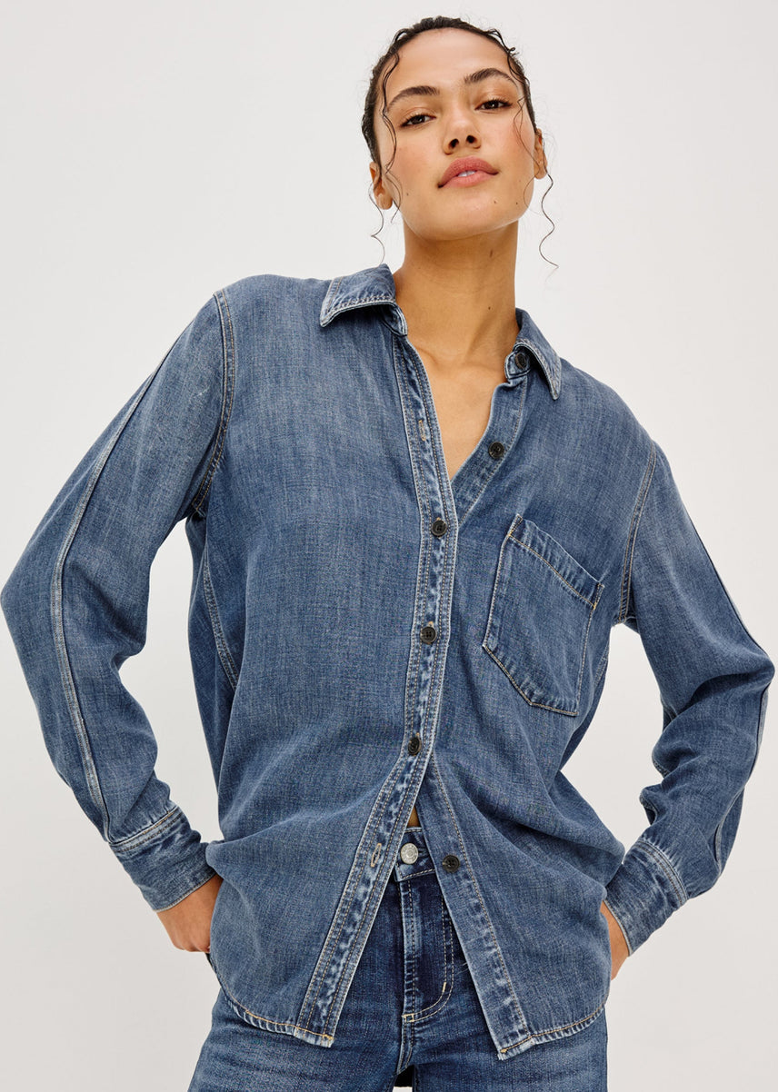 A woman wearing a blue denim button-up shirt with a chest pocket, paired with matching blue jeans, photographed against a plain light gray studio background.