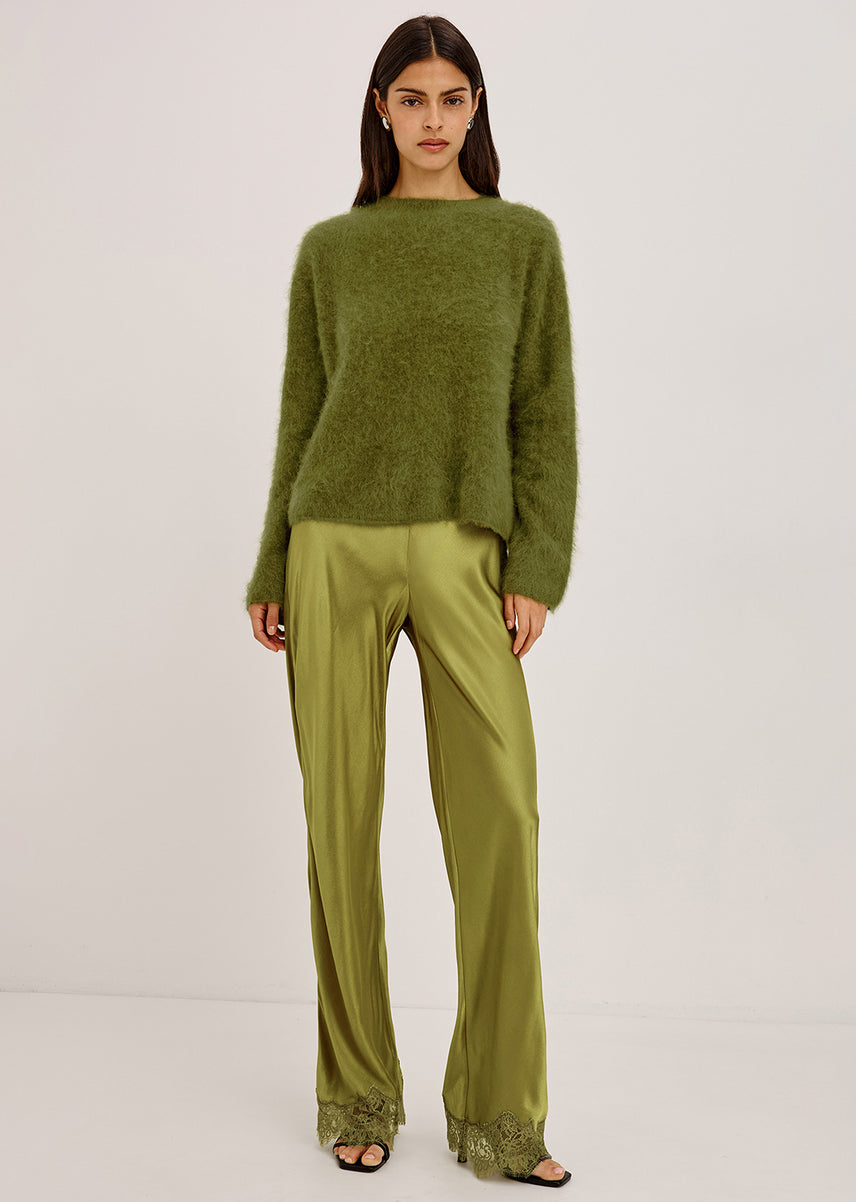 A model stands against a plain light gray studio background wearing an olive green fuzzy long-sleeve sweater and matching olive green satin wide-leg trousers with lace trim at the hems.