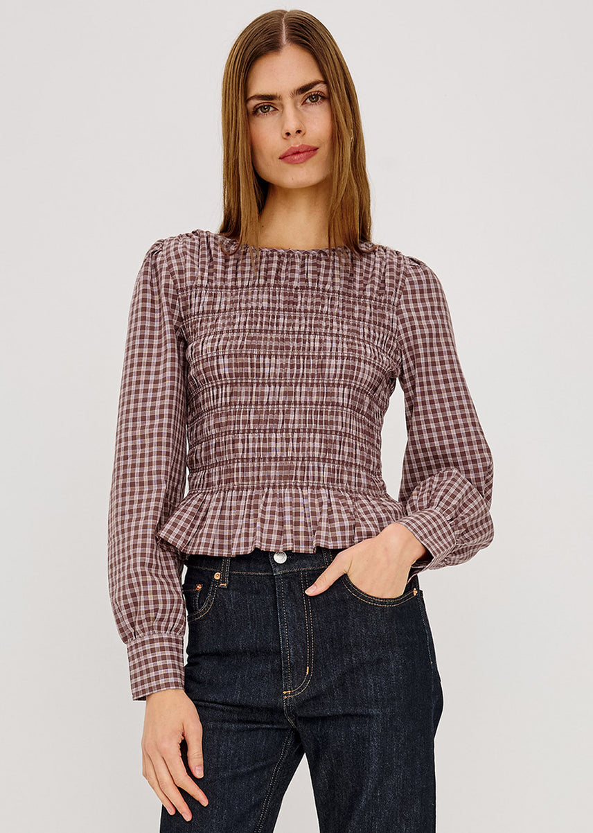 A woman modeling a long-sleeve smocked blouse with a brown and beige gingham pattern, paired with dark blue jeans, against a light gray studio background.