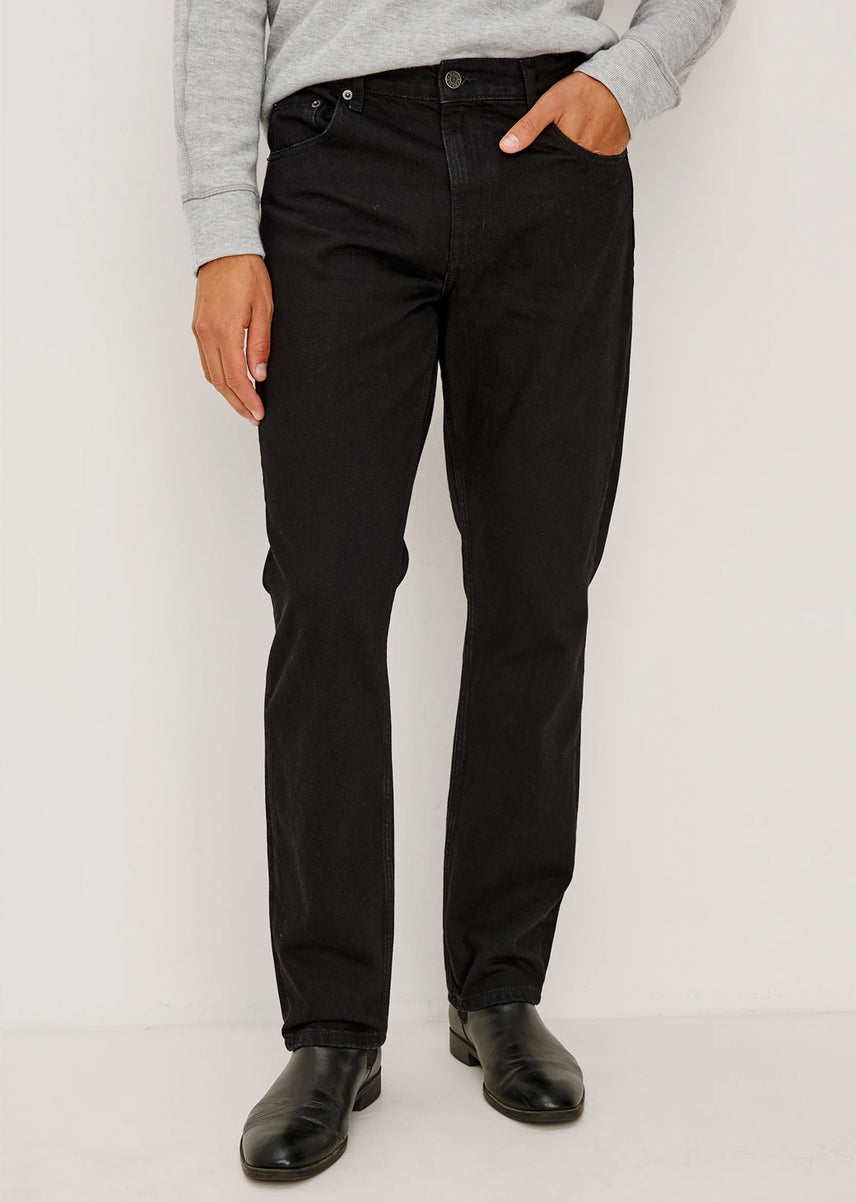 A person wearing black slim-fit jeans with a light gray long-sleeve top and black leather shoes, standing against a plain white studio background.