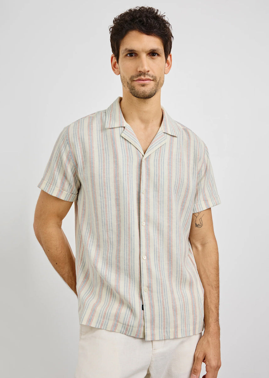 A man wearing a short-sleeve button-up shirt with vertical stripes in beige, white, and soft pastel colors, paired with off-white trousers, against a light gray studio background.