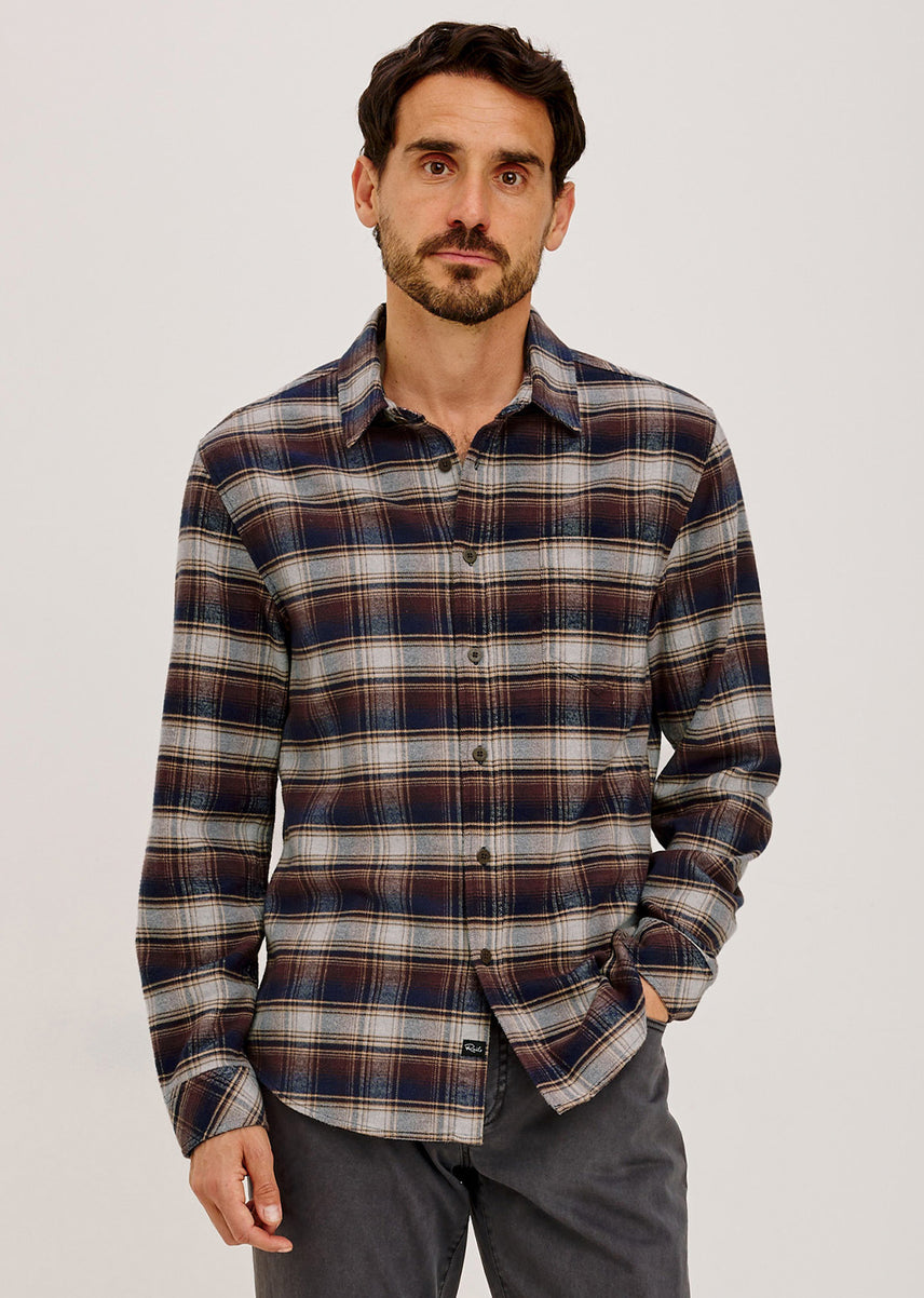 A man wearing a long-sleeve button-up plaid shirt in navy, brown, and beige tones, paired with dark gray trousers, photographed against a light gray studio background.