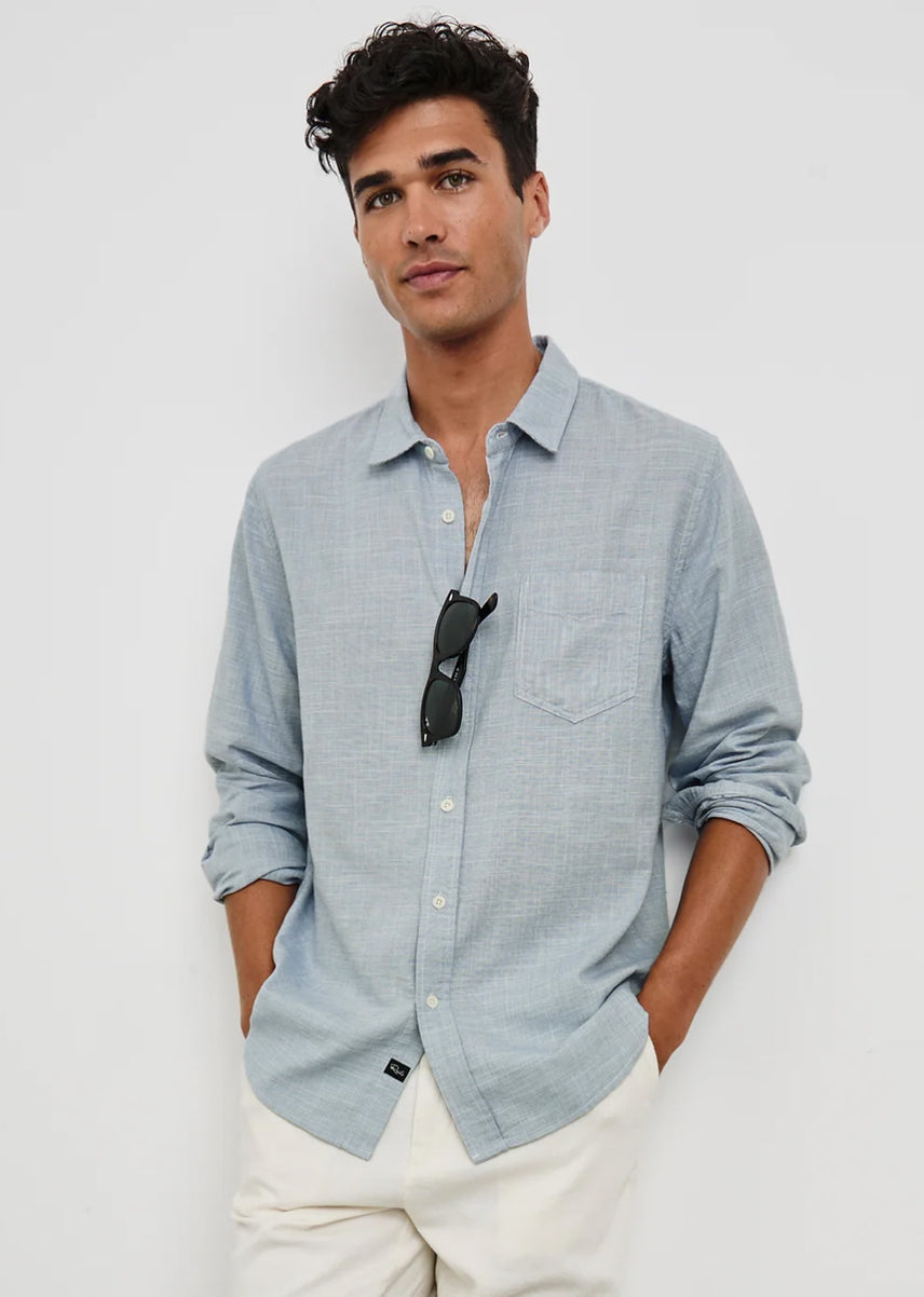 A man wearing a light blue long-sleeve linen button-down shirt with white trousers, standing against a plain white studio background with sunglasses hanging from his shirt collar.