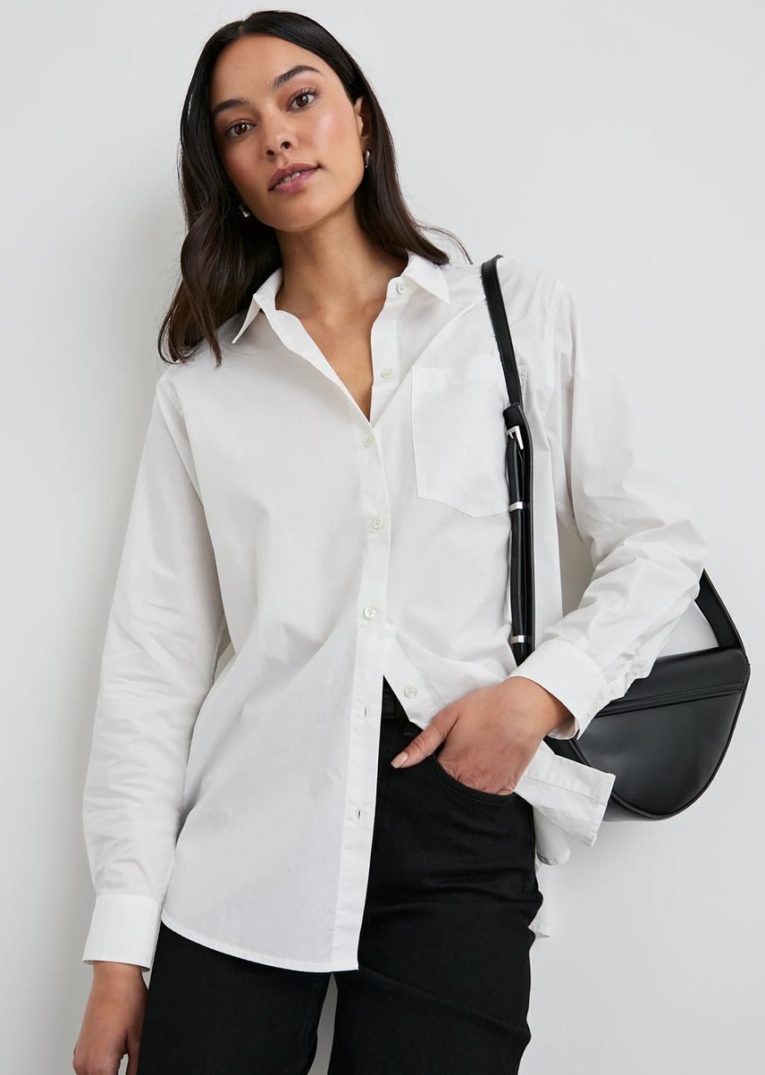 Woman wearing a white shirt and black pants with a black handbag against a plain background
