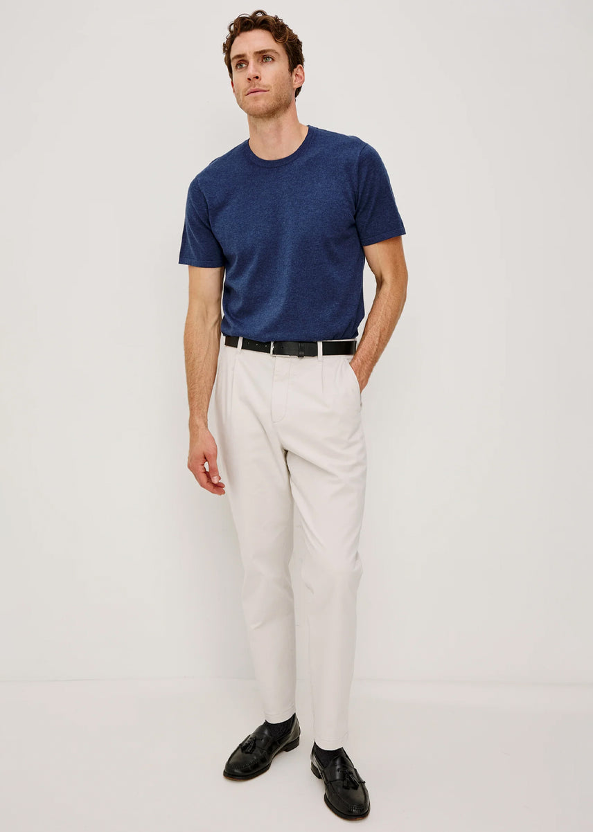 A man modeling a navy blue short-sleeve crewneck t-shirt, off-white trousers, a black belt, and black leather shoes, standing against a plain light gray studio background.