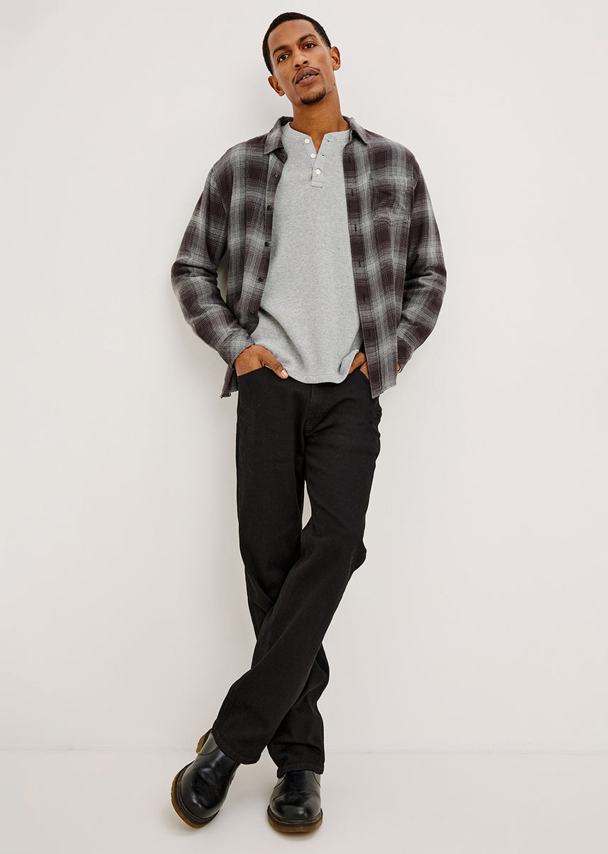 A man modeling a casual outfit consisting of a gray and black plaid button-down shirt over a light gray henley t-shirt, paired with black trousers and black leather shoes, standing against a plain white studio background.