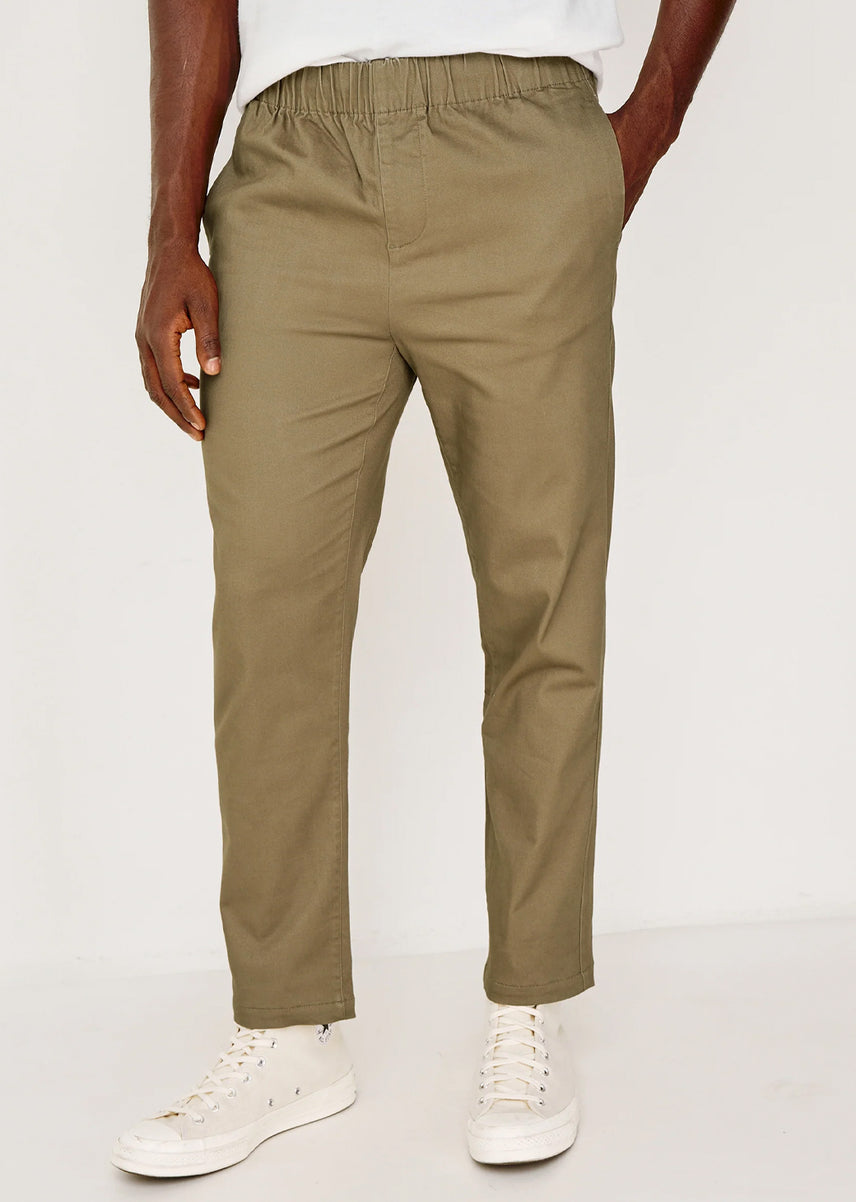A person wearing olive green chinos with a white t-shirt and white sneakers, shown from the waist down against a plain white studio background.