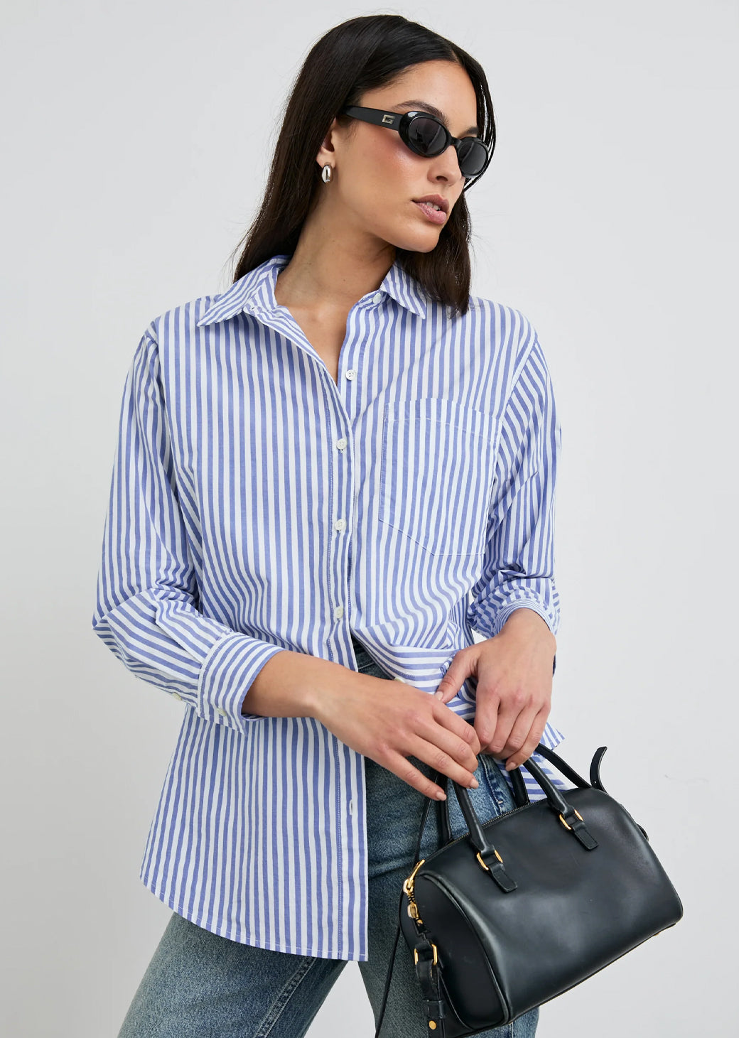 Woman wearing a blue and white striped button down shirt holding a black handbag against a gray background