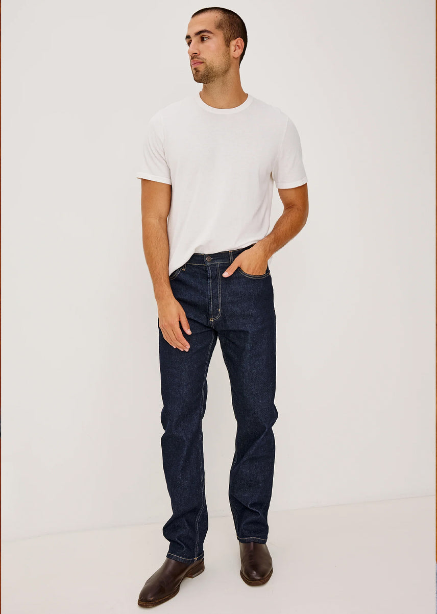A man wearing a white crew-neck t-shirt, dark blue straight-leg jeans, and brown leather shoes, standing against a plain off-white studio background.