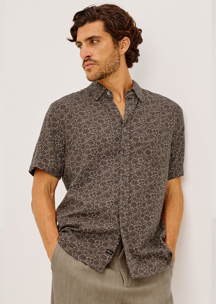 A man wearing a short-sleeve button-up shirt with a dark brown abstract pattern, paired with trousers, standing against a plain light gray background.