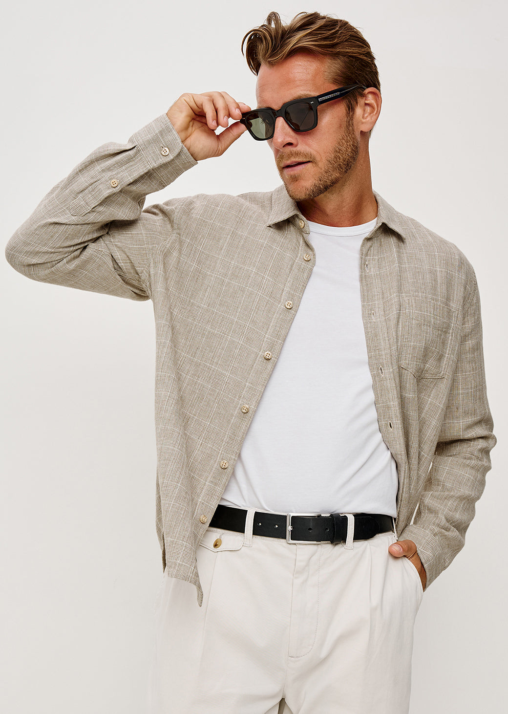 Man wearing a beige shirt over a white t-shirt with sunglasses on a plain background