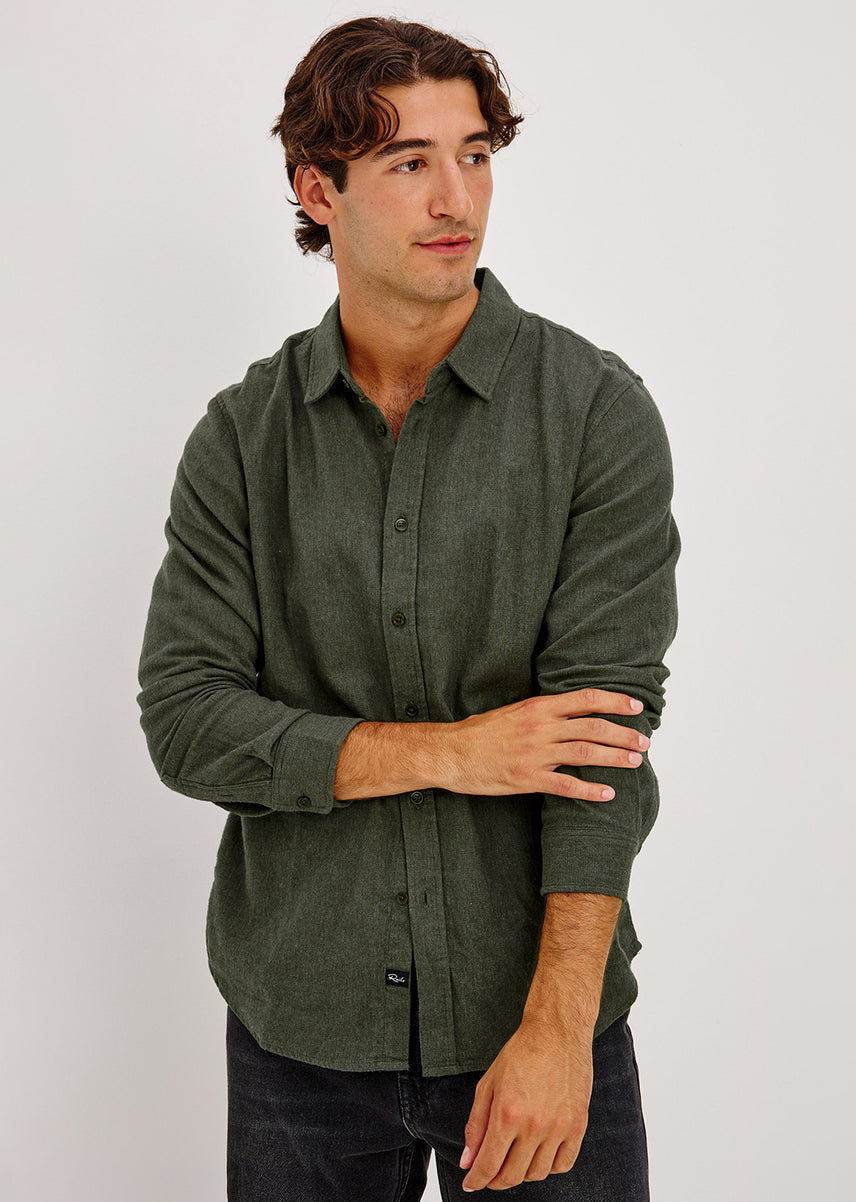 A man wearing a dark olive green long-sleeve button-up shirt, paired with dark jeans, standing against a plain light gray studio background.