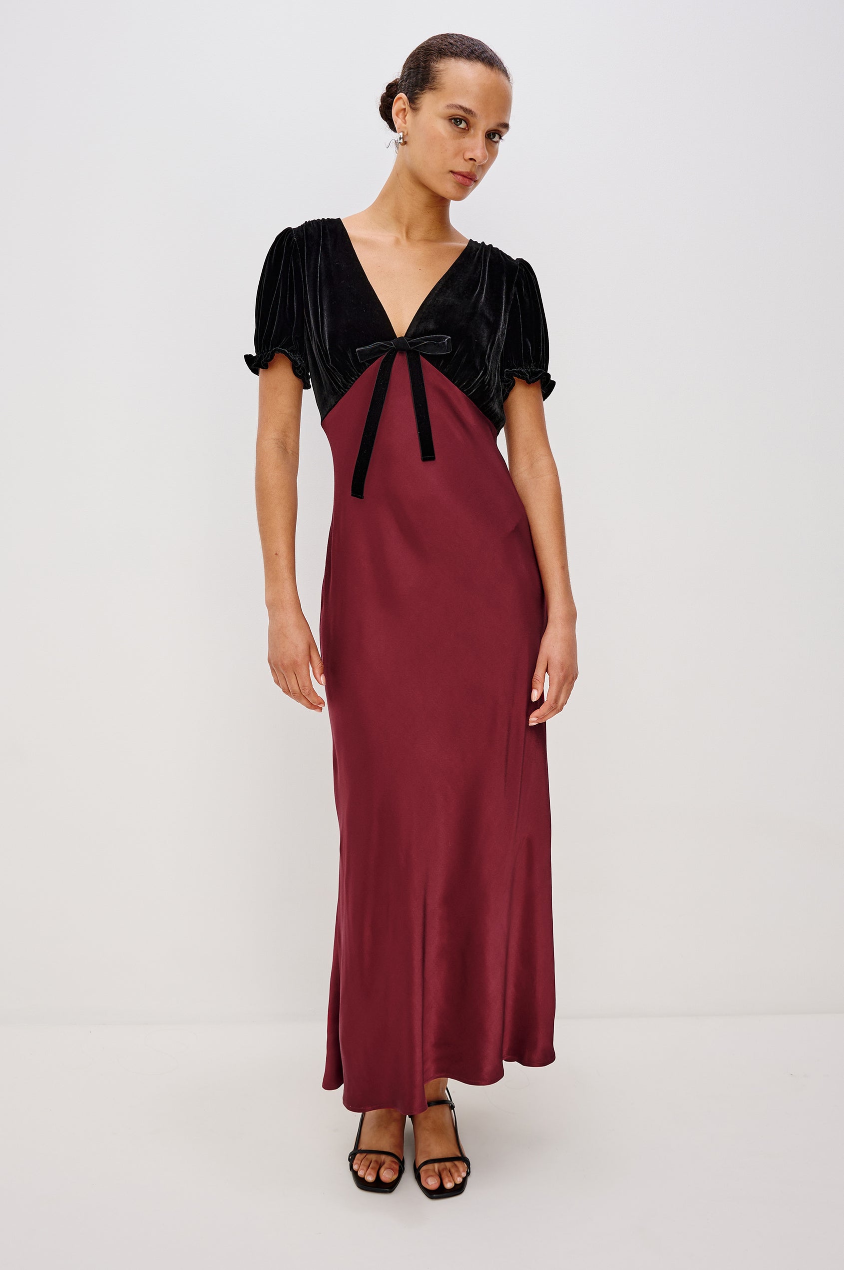 A garnet velvet maxi dress with a fitted V-neck bodice and adjustable center-bust ties. The bias-cut skirt drapes elegantly to ankle length, finished with a back zipper closure. Front View on Model.