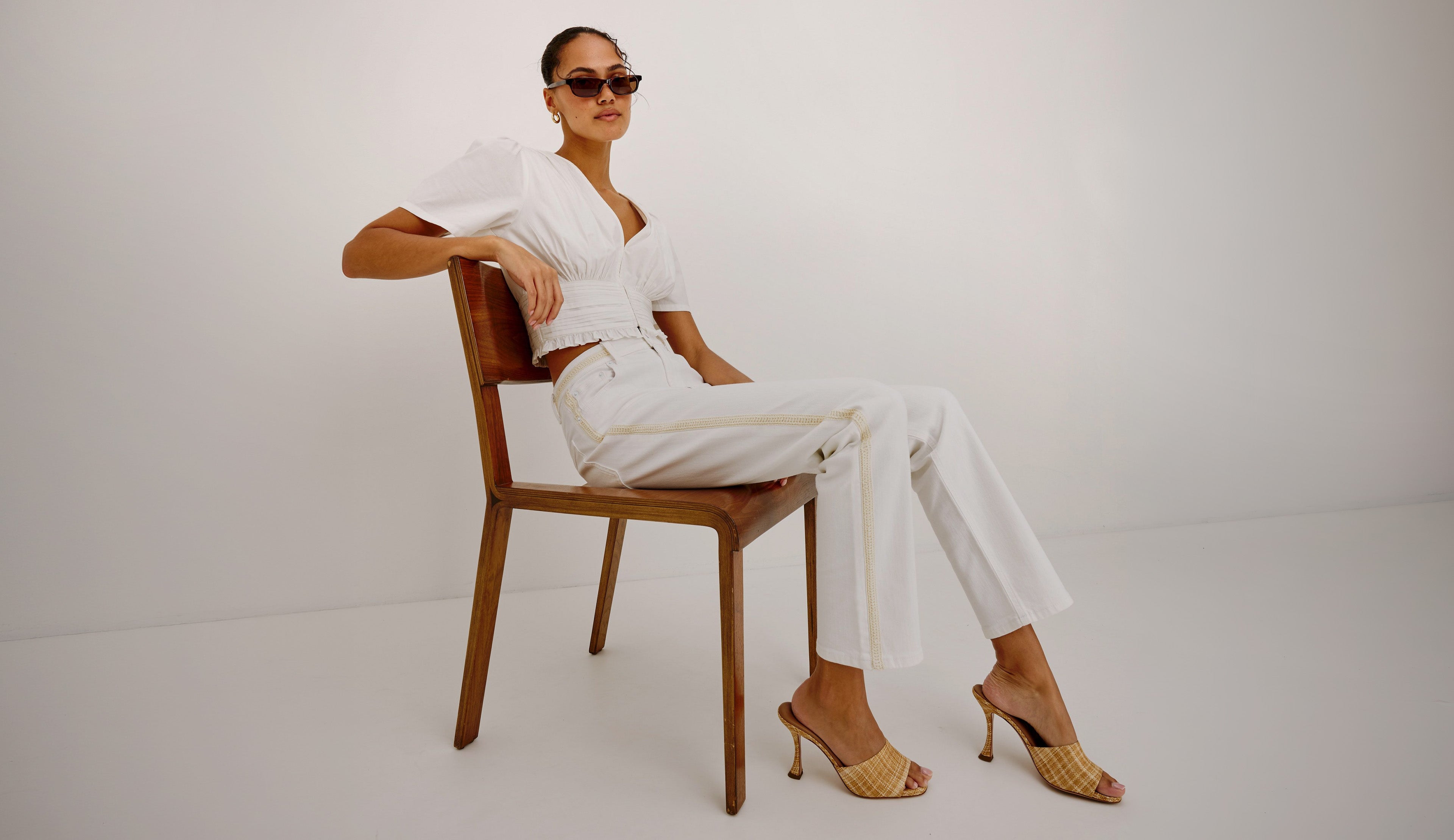 A woman wearing a white short-sleeve top and white wide-leg trousers sits on a wooden chair against a plain light gray studio background. She is wearing gold heeled sandals and dark sunglasses.
