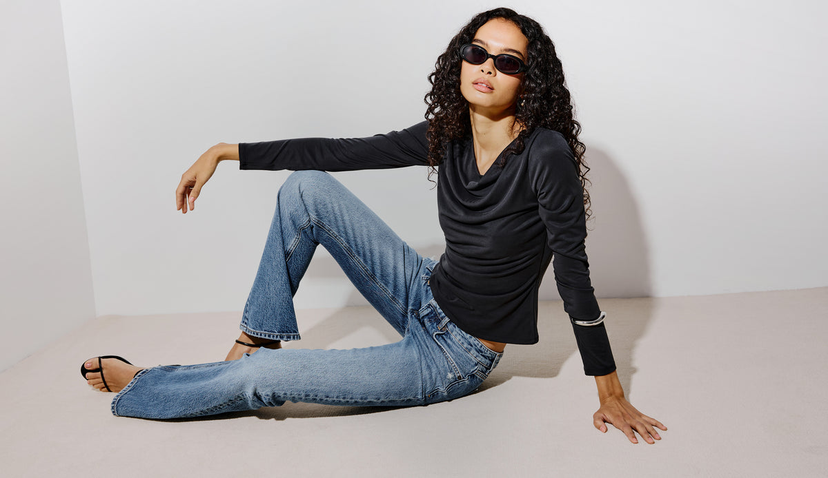 A woman wearing a black long-sleeve cowl-neck top and light-wash blue jeans sits on a minimalist light gray floor against a matching light gray wall.