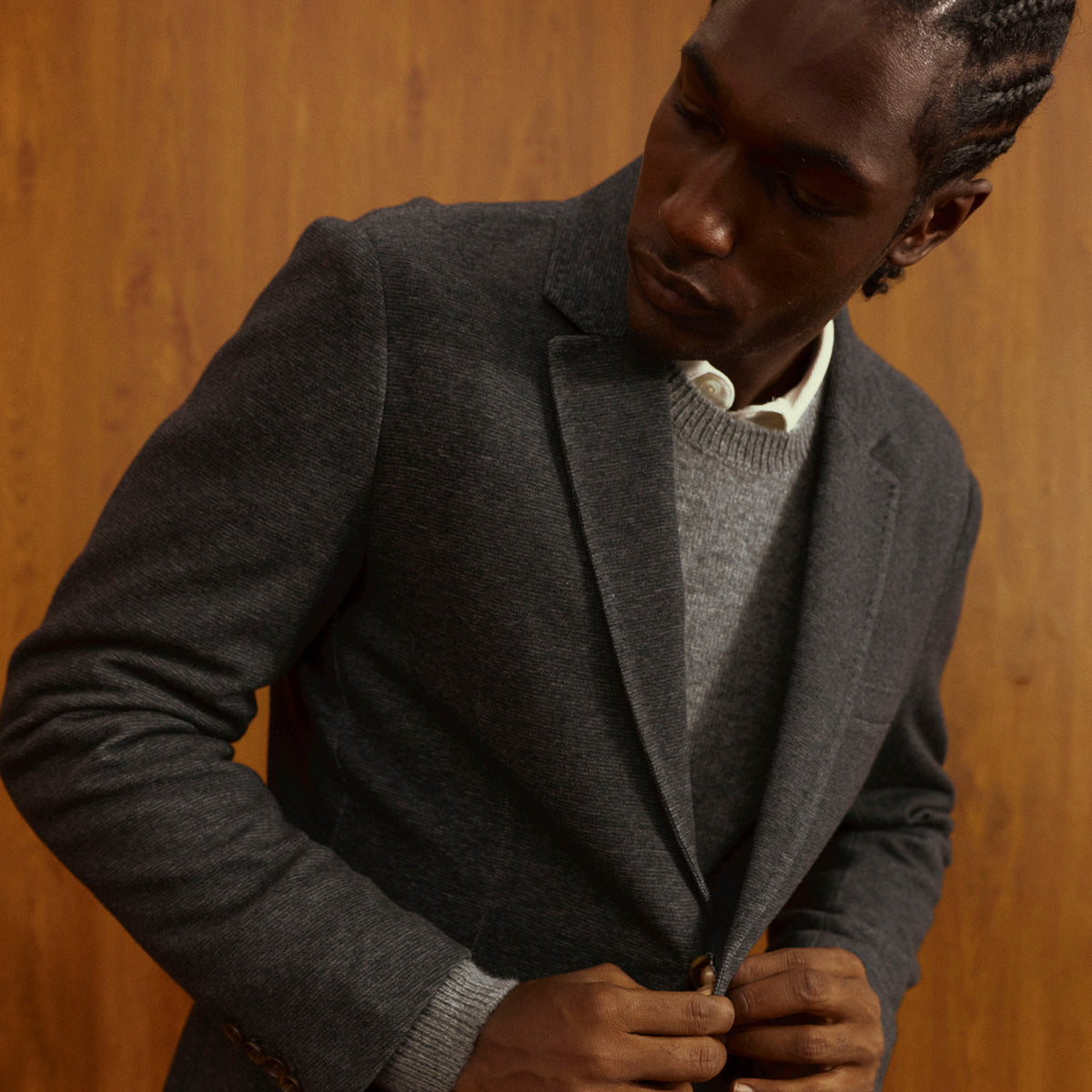 Man in a gray blazer over a gray crewneck sweater and white collared shirt, against a warm wood-paneled wall.